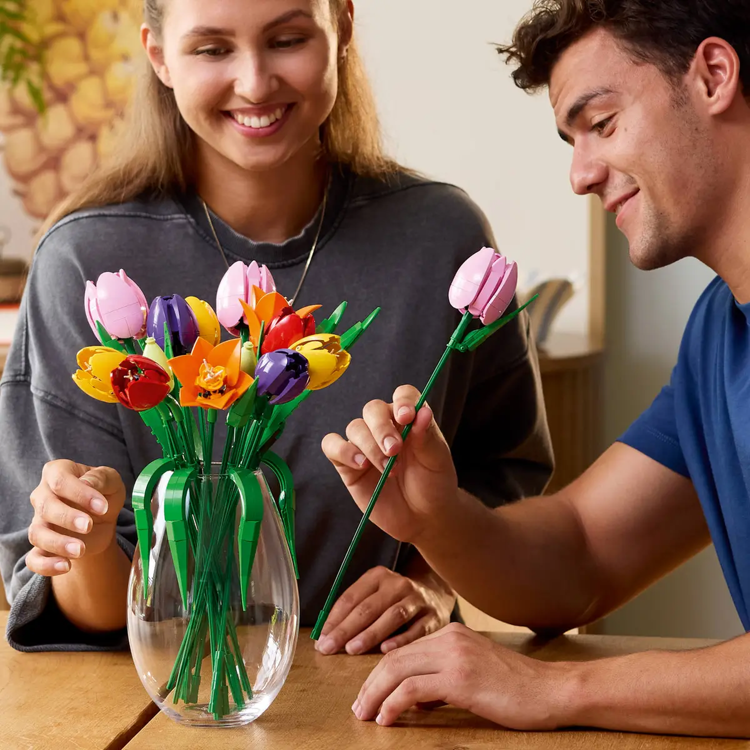 Een stel glimlacht naar een vaas met kleurrijke Lego-tulpen. Man houdt een roze tulp vast. De vaas staat op een houten tafel.