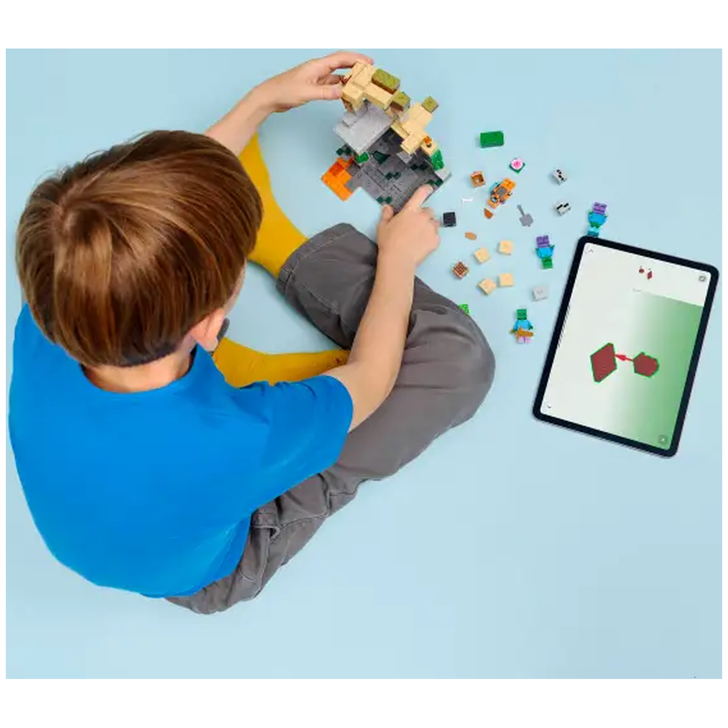 Een kind bouwt met Lego-stenen, volgens instructies op een tablet. Verschillende Lego-onderdelen en figuren liggen verspreid.