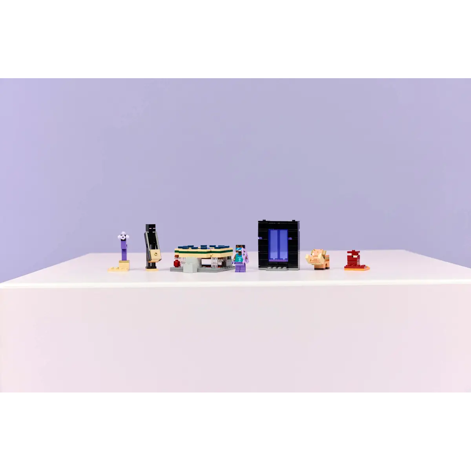 A white table holds various Minecraft Lego sets: a portal, figures, and structures against a purple backdrop.
