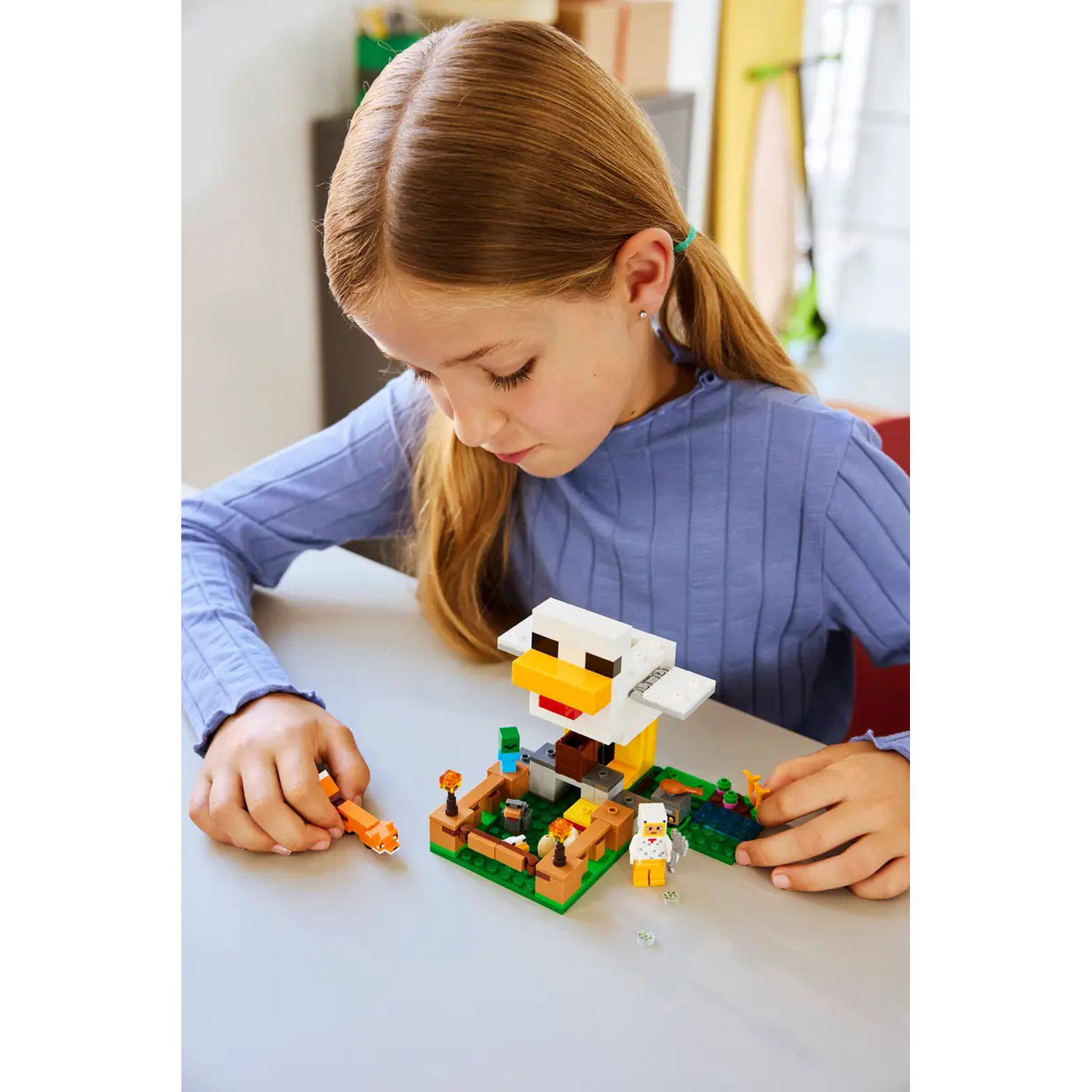 Een meisje speelt met een Minecraft Lego-set op een tafel. De set bevat een kippenhok, personages en andere Legoblokken.