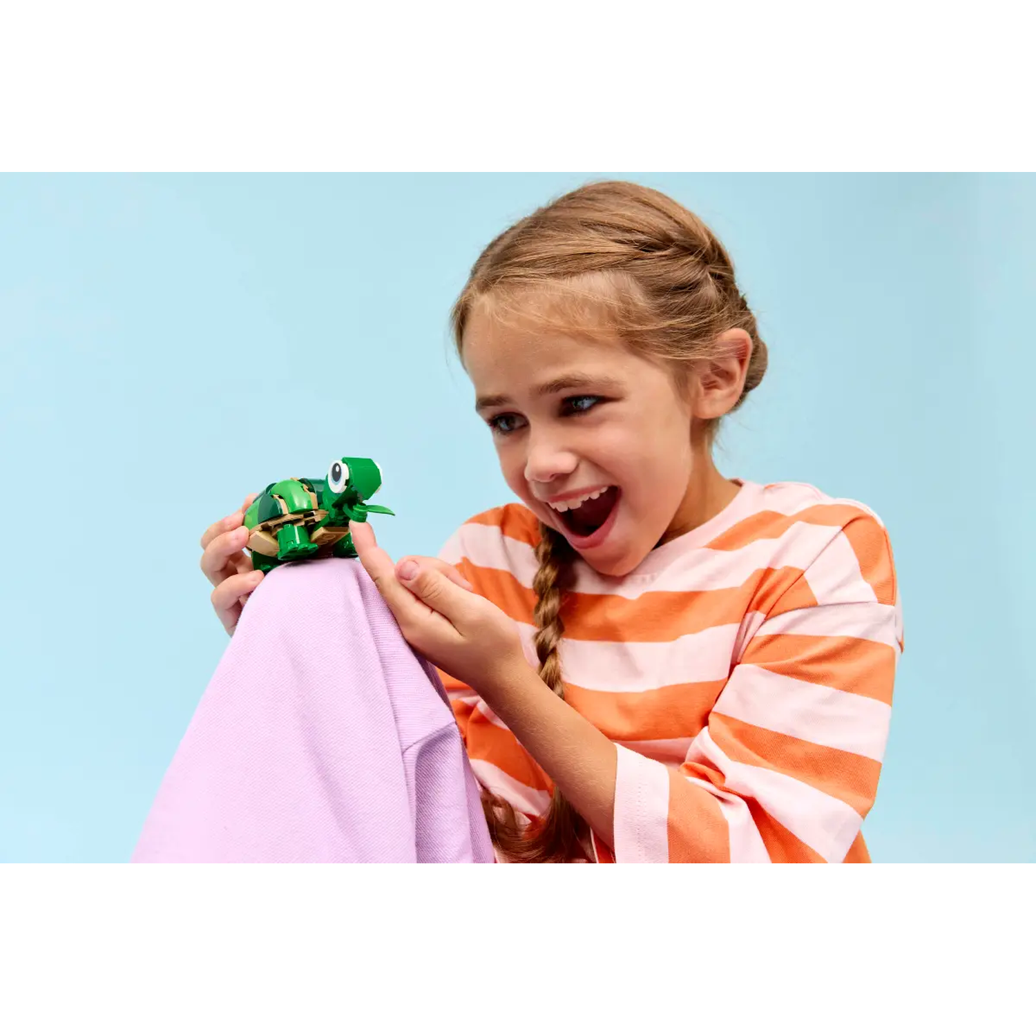 A girl with a braid smiles, holding a green Lego turtle. She wears an orange striped shirt. A purple cloth is in the foreground.