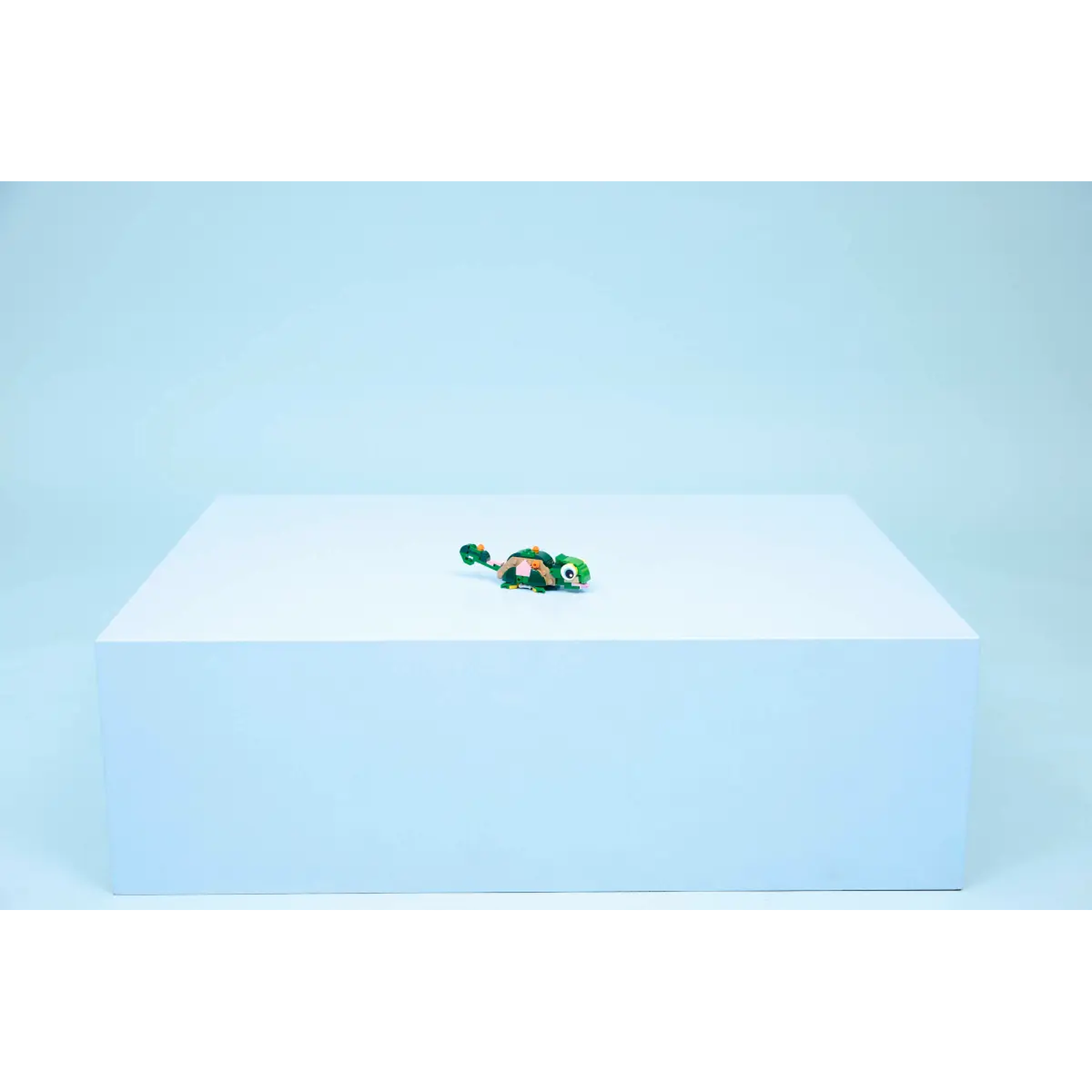 A small toy turtle on a white block against a light blue background. The turtle is green, orange, and white.
