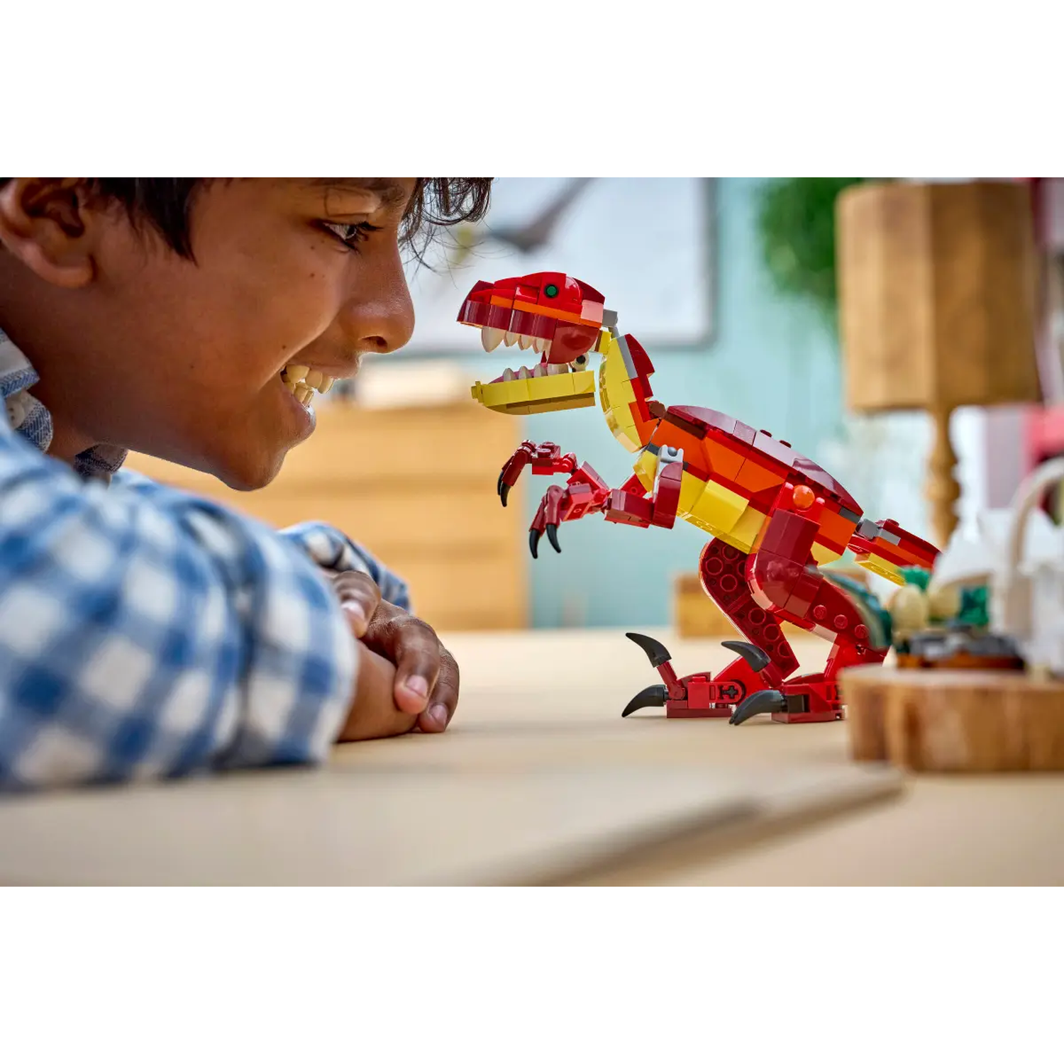 A boy smiles at a Lego dinosaur, red, yellow, and orange. The dinosaur is on a wooden surface.