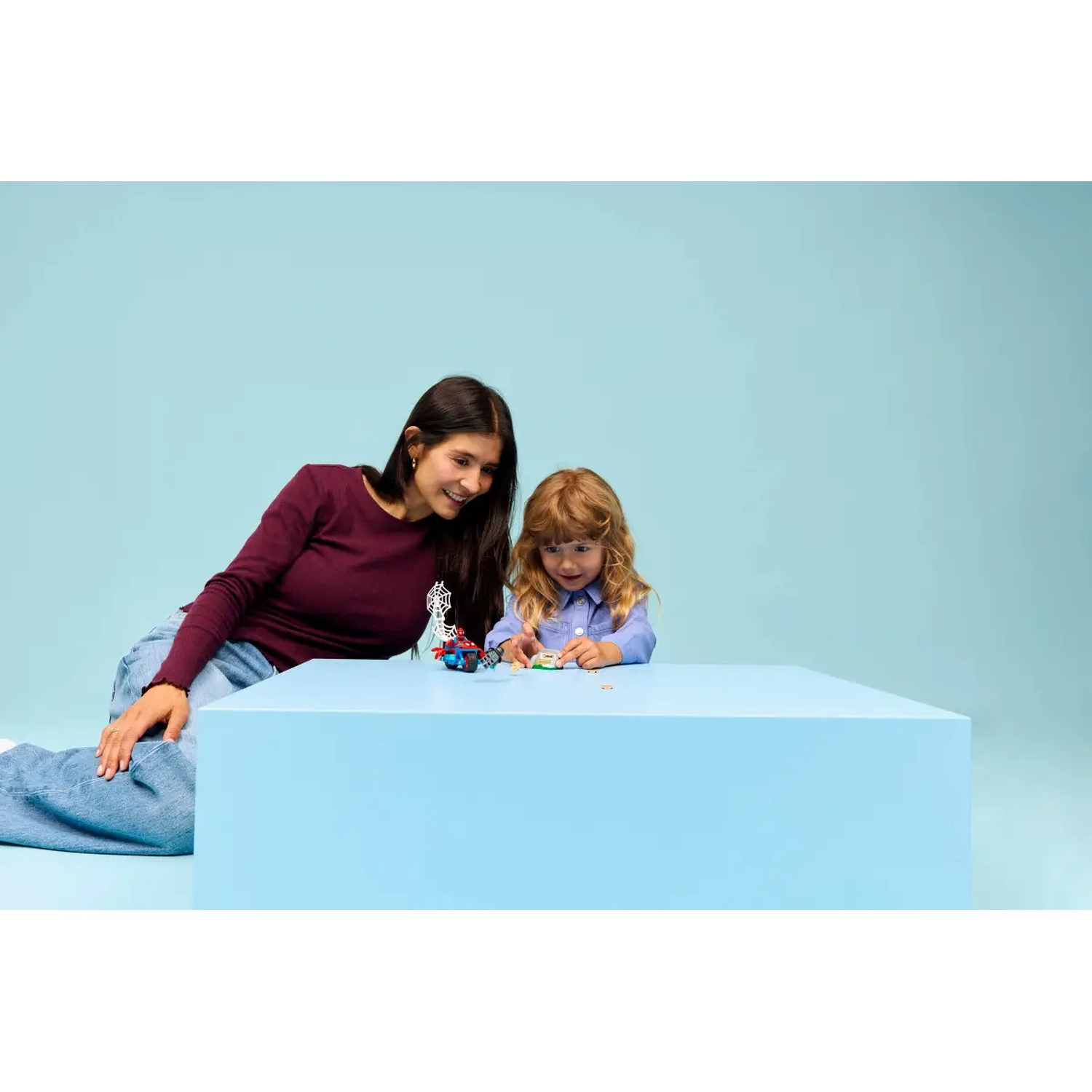 Een vrouw en een kind spelen met speelgoed op een blauwe tafel. De vrouw glimlacht. De achtergrond is ook blauw.