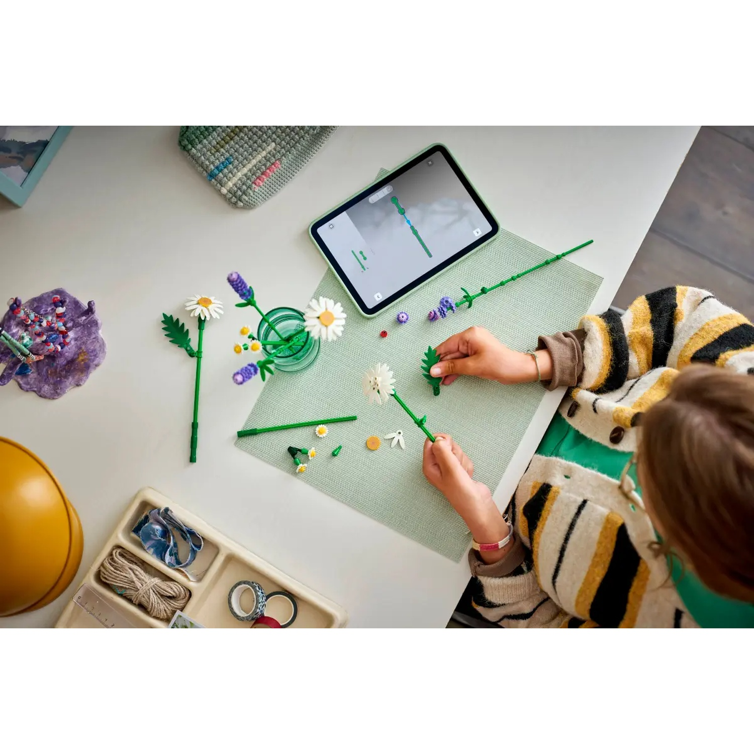 Une personne assemble des modèles de fleurs sur une table blanche. Un iPad et du matériel de bricolage sont présents. La personne porte un pull rayé.
