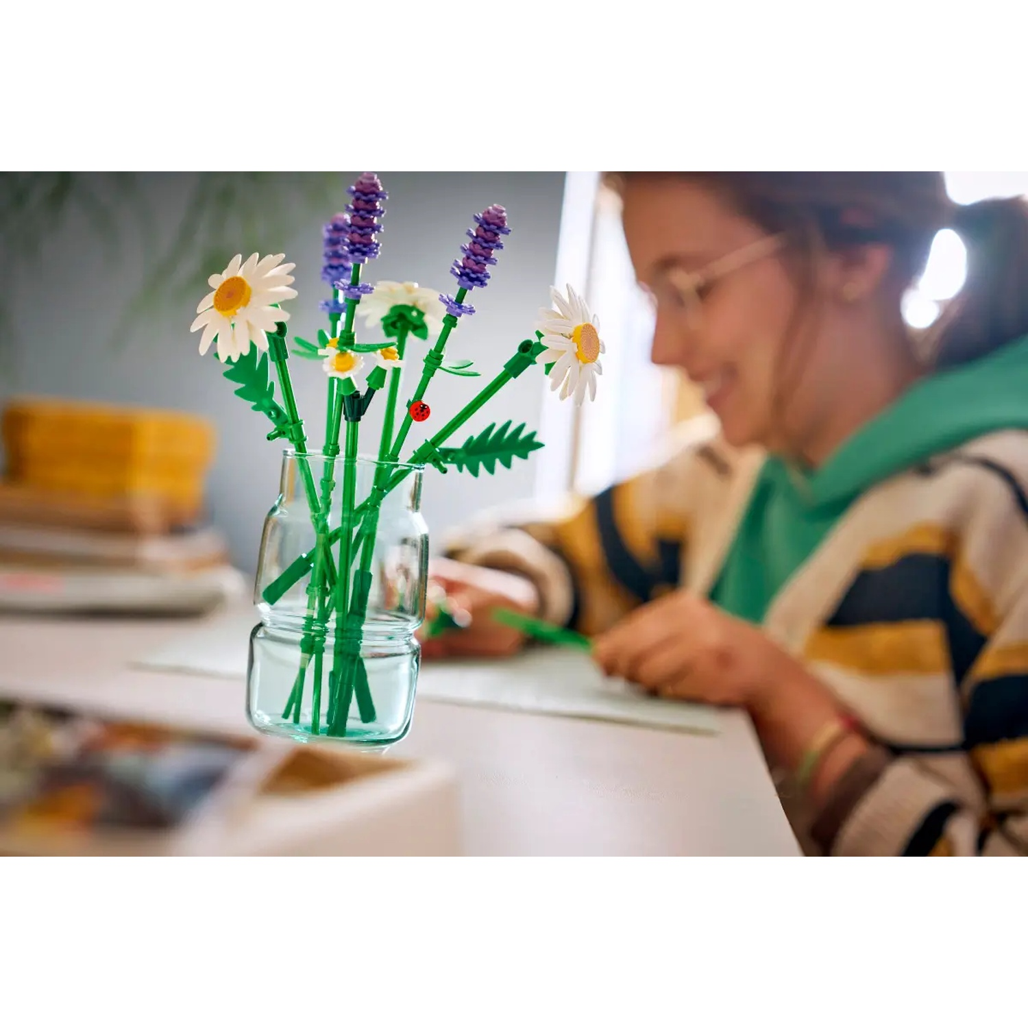 Un vase transparent avec des fleurs Lego. Une fille avec des lunettes écrit à un bureau en arrière-plan. Des marguerites et de la lavande sont dans le vase.