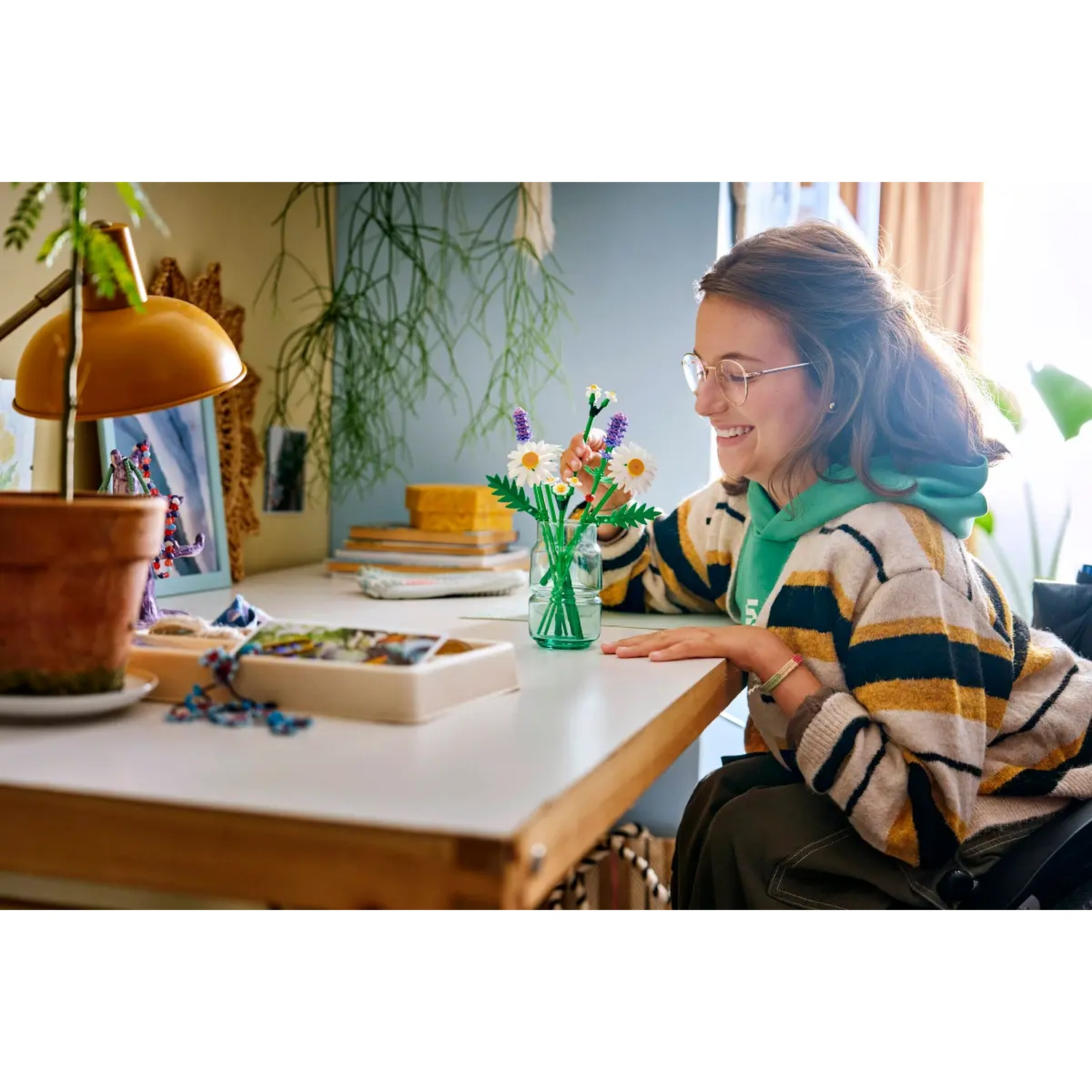 Une jeune fille souriante avec des lunettes arrange des fleurs jouets sur un bureau blanc. Une lampe de bureau et des plantes sont en arrière-plan.