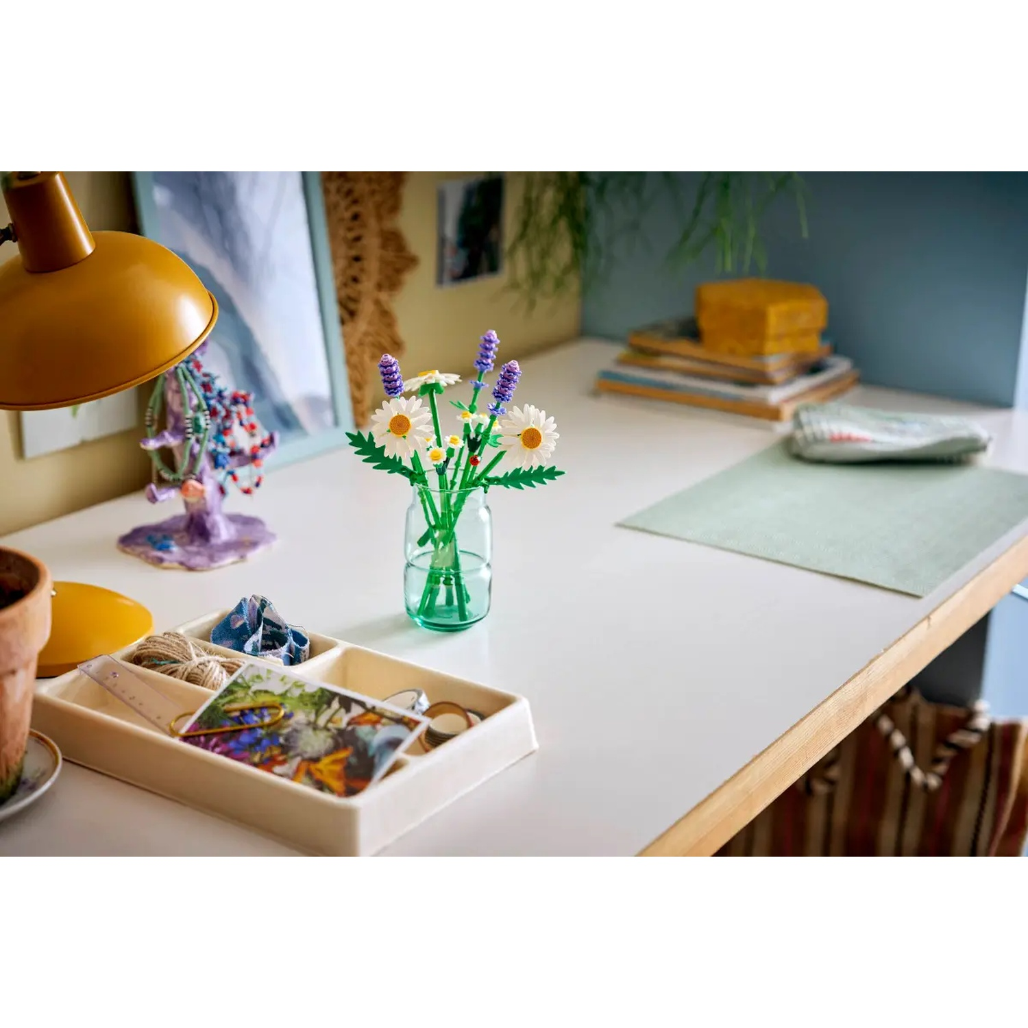 Un bureau blanc avec un vase à fleurs, une lampe et un plateau avec des objets. Au fond, une pile de livres et un tableau encadré.