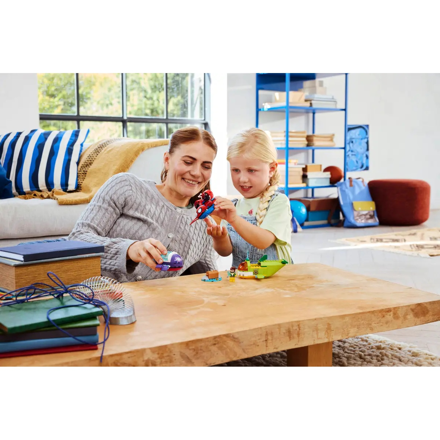 Une femme et une fille jouent avec des jouets LEGO sur une table en bois. Un canapé, des étagères et une fenêtre sont en arrière-plan.