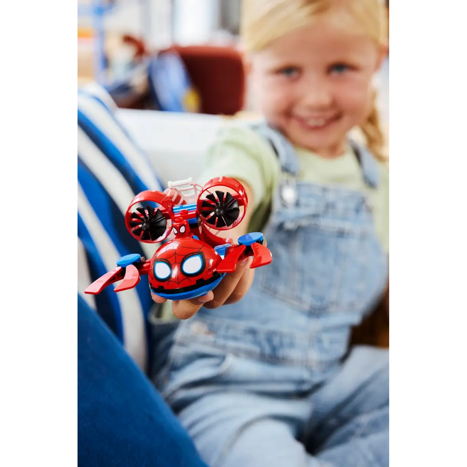 Une fille tient un jouet Spider-Man. Le jouet est rouge, bleu et noir. Elle porte une salopette en jean et sourit.