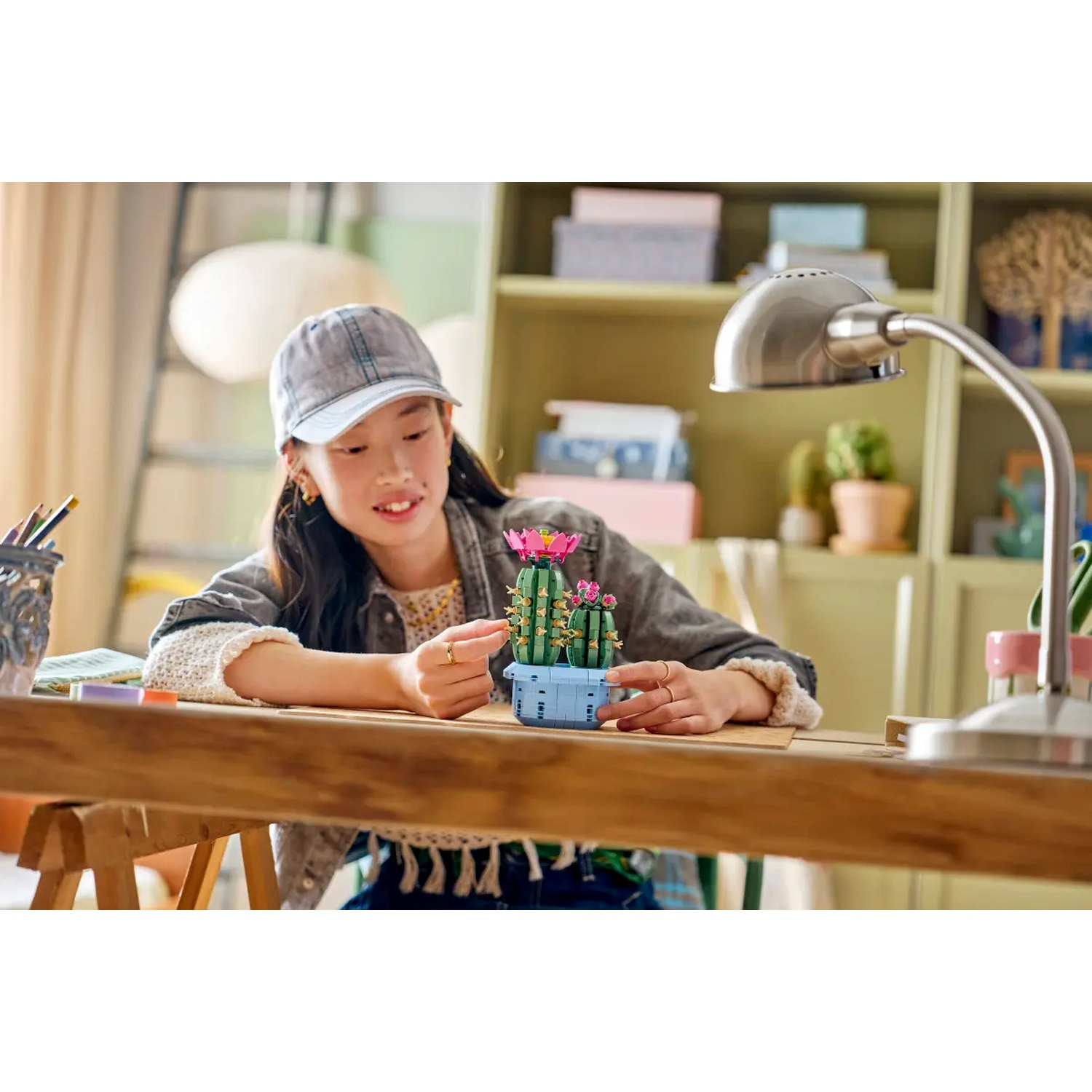 Une jeune femme avec une casquette arrange un cactus Lego sur un bureau en bois. Une lampe, des étagères et une échelle sont en arrière-plan.