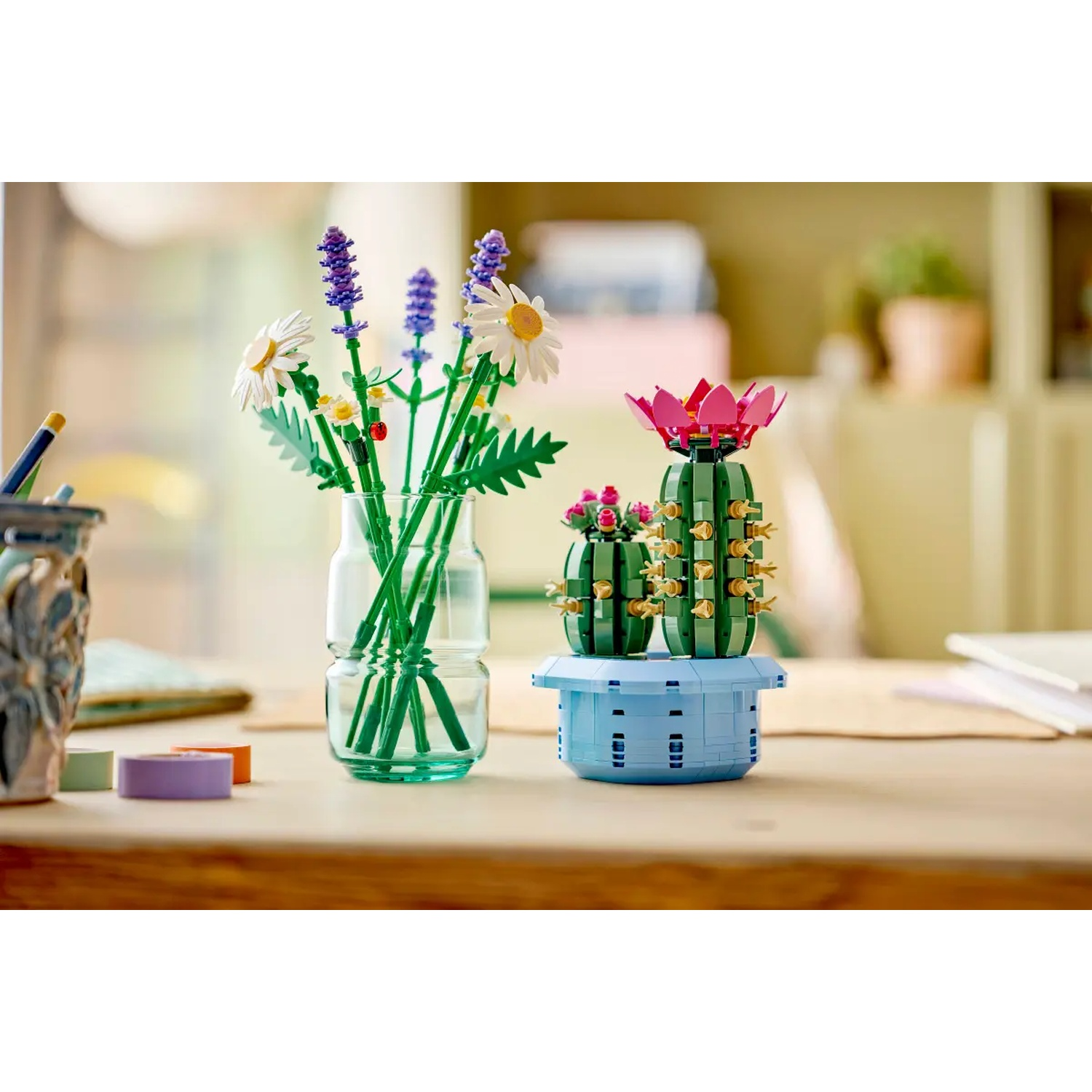 Une table présente des fleurs et un cactus Lego. Les fleurs sont dans un vase en verre et le cactus dans un pot bleu. Le fond est flou.