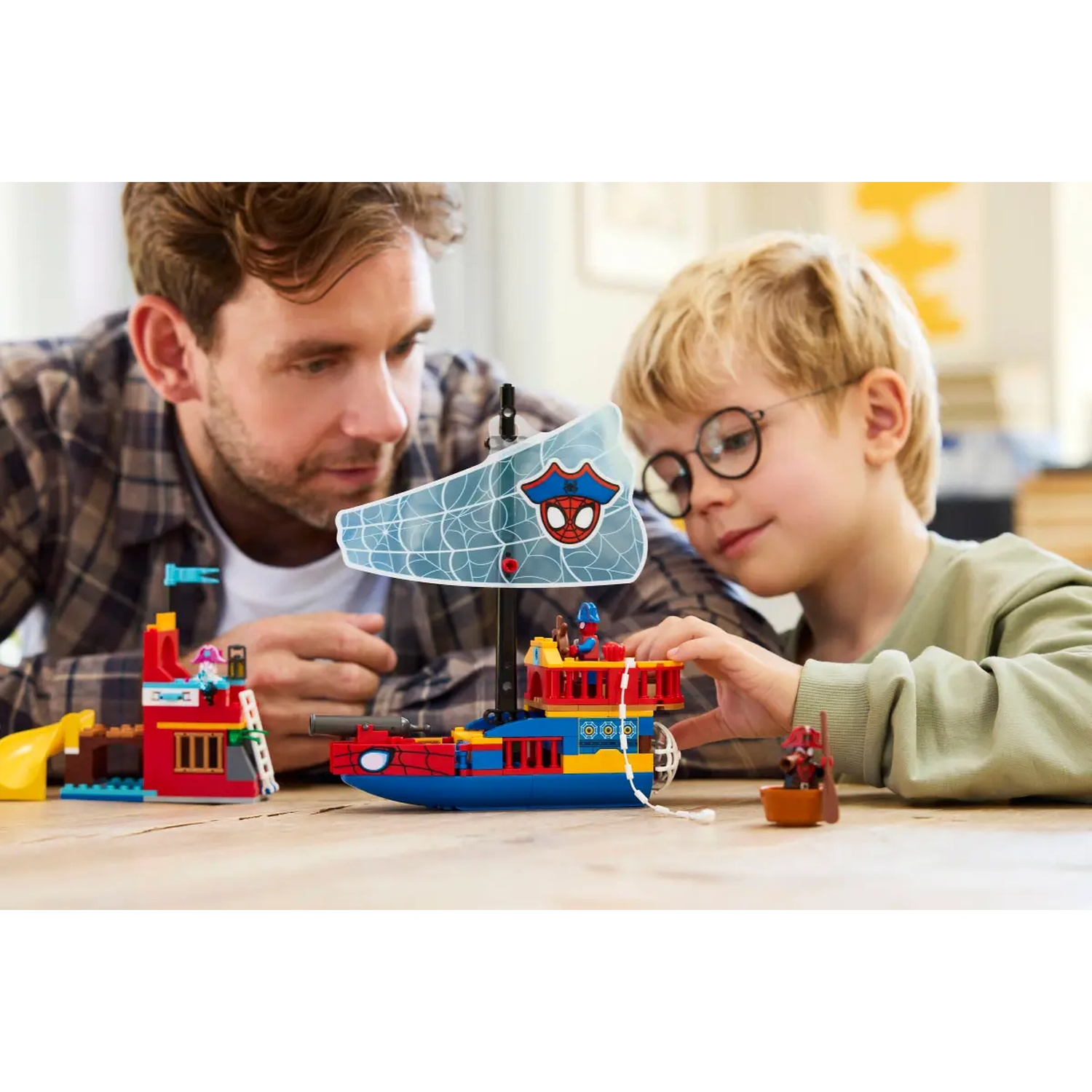 A man and a boy are playing with a Lego Spiderman boat. The boat has a Spiderman sail. A building is on the left.