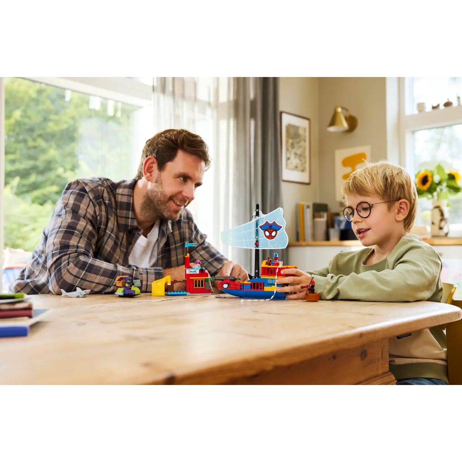 A man and a boy play with toy boats on a wooden table. The scene is indoors, with a window and a vase of flowers visible.