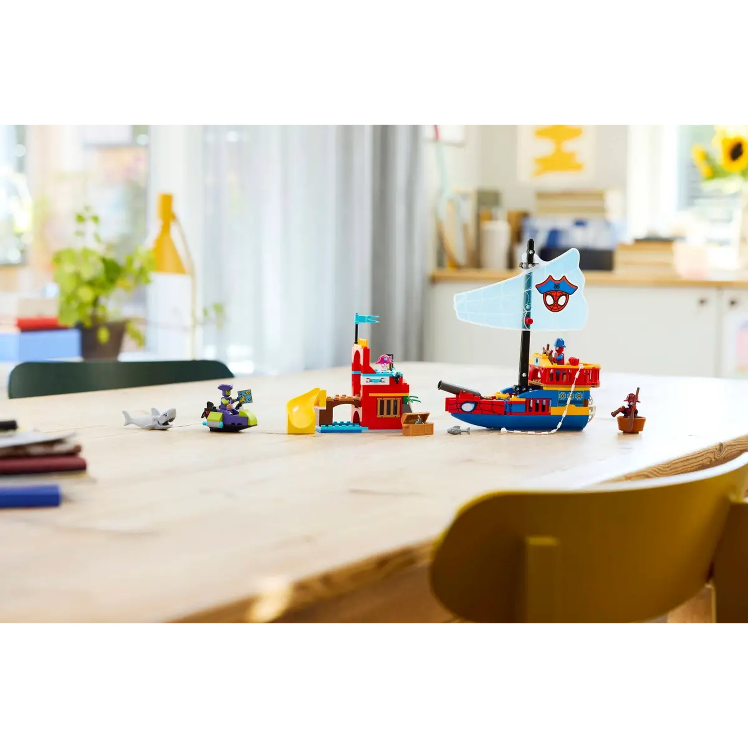 A table with Lego sets of a pirate ship, a small island, and characters like Spider-Man and a shark. A yellow chair is in the foreground.