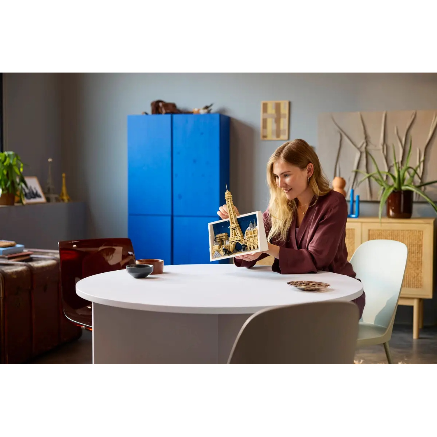 Woman holding a model of the Eiffel Tower, sitting at a white table. A blue cabinet is in the background.