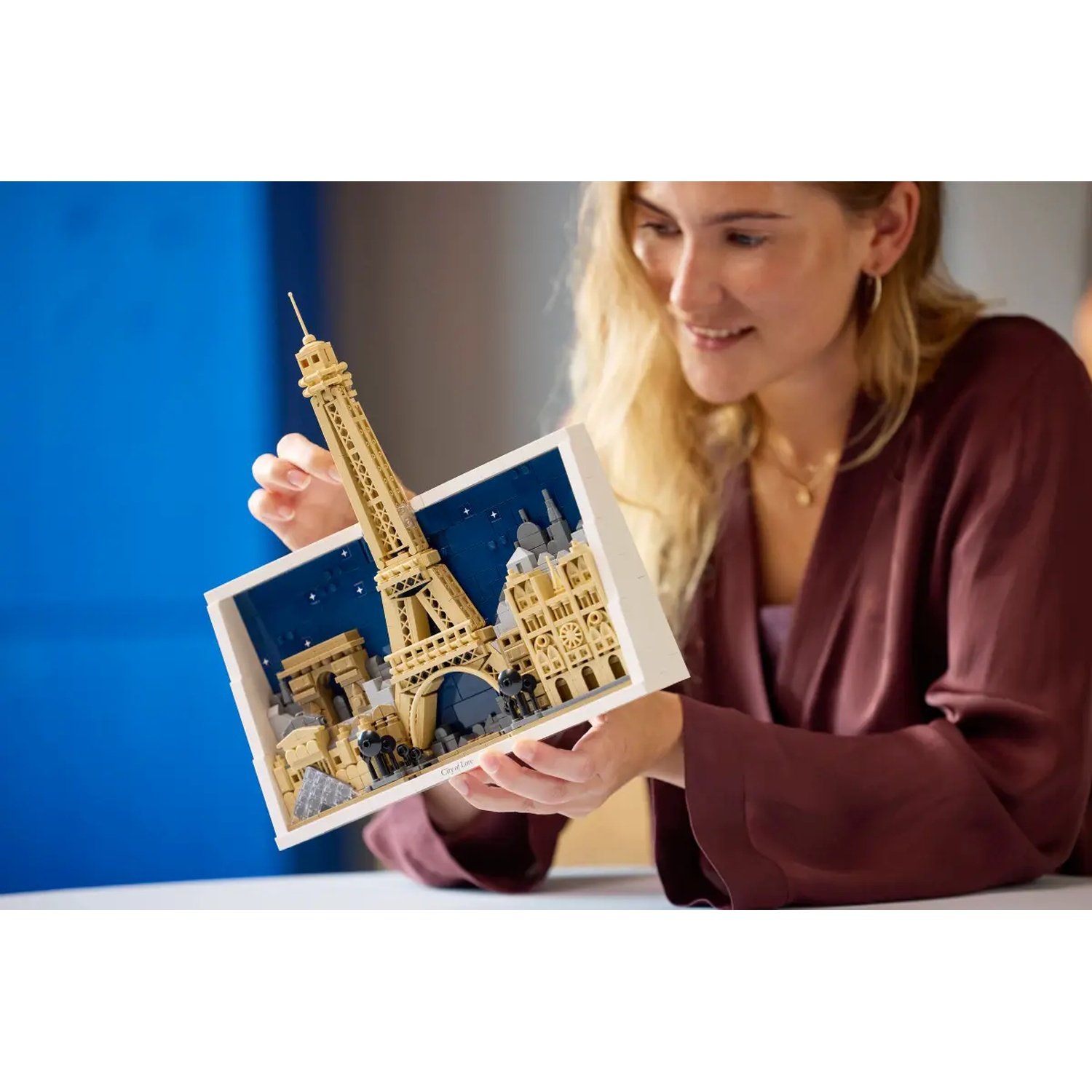 A woman holds a Lego diorama of Paris, including the Eiffel Tower. She is smiling. The background is blurred.