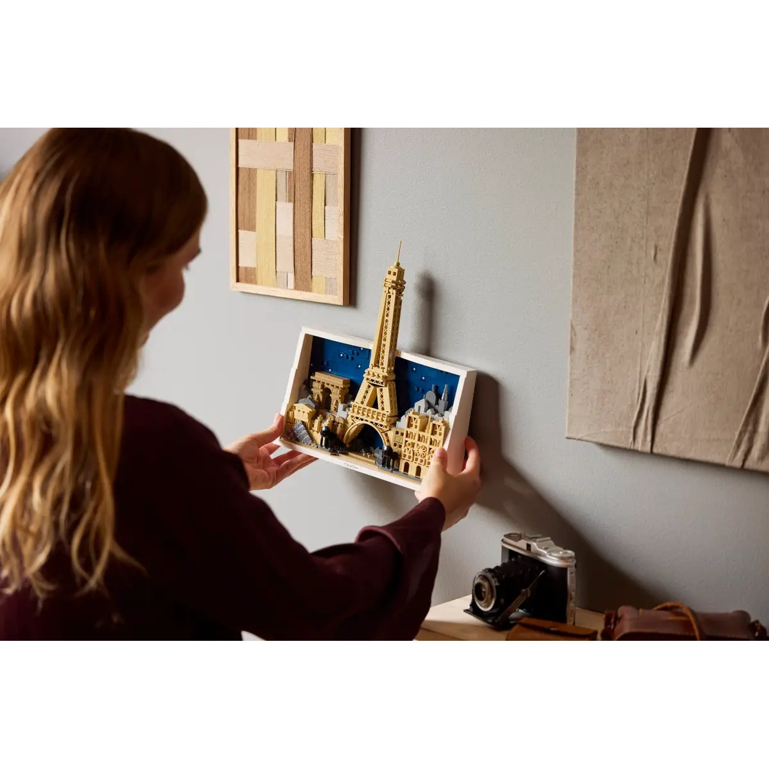 A woman holds a Lego Eiffel Tower art piece. Background includes framed art, a camera, and a leather bag on a wooden surface.