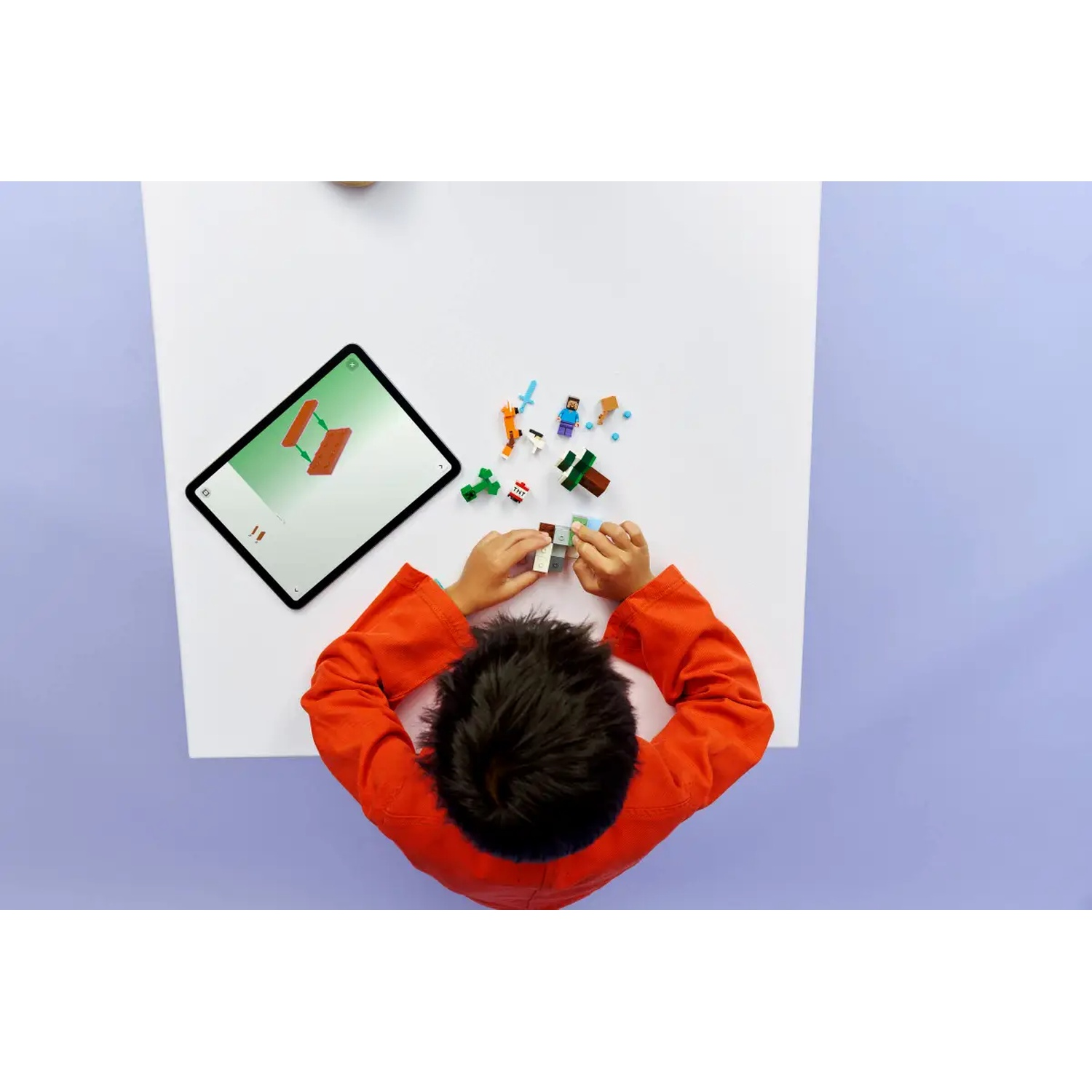 Un enfant en chemise orange assemble des pièces Lego sur une table blanche. Une tablette affiche des instructions. Diverses figurines et blocs Lego sont éparpillés.