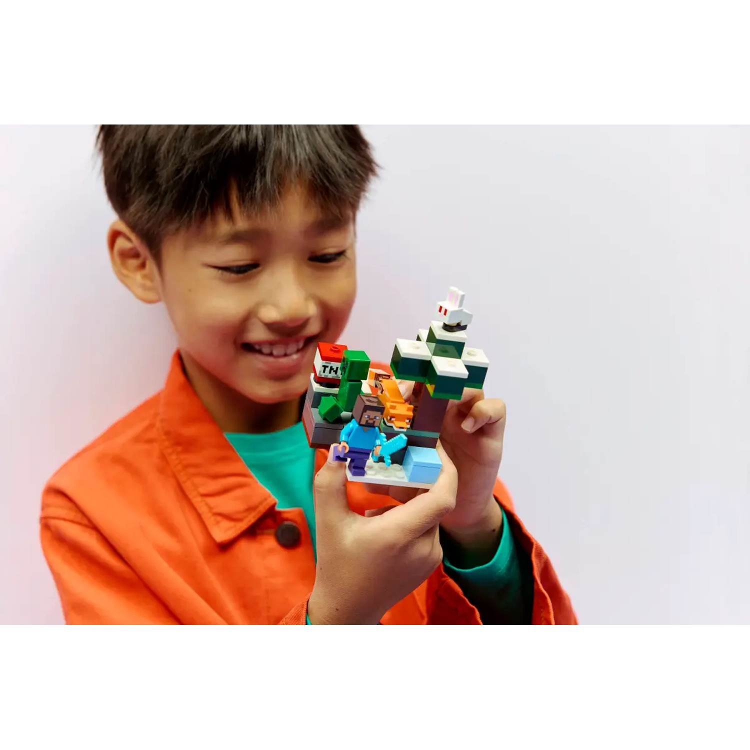Un garçon souriant tient un set Lego Minecraft. L'ensemble montre un personnage, un renard et un arbre. Le garçon porte une veste orange et une chemise verte.