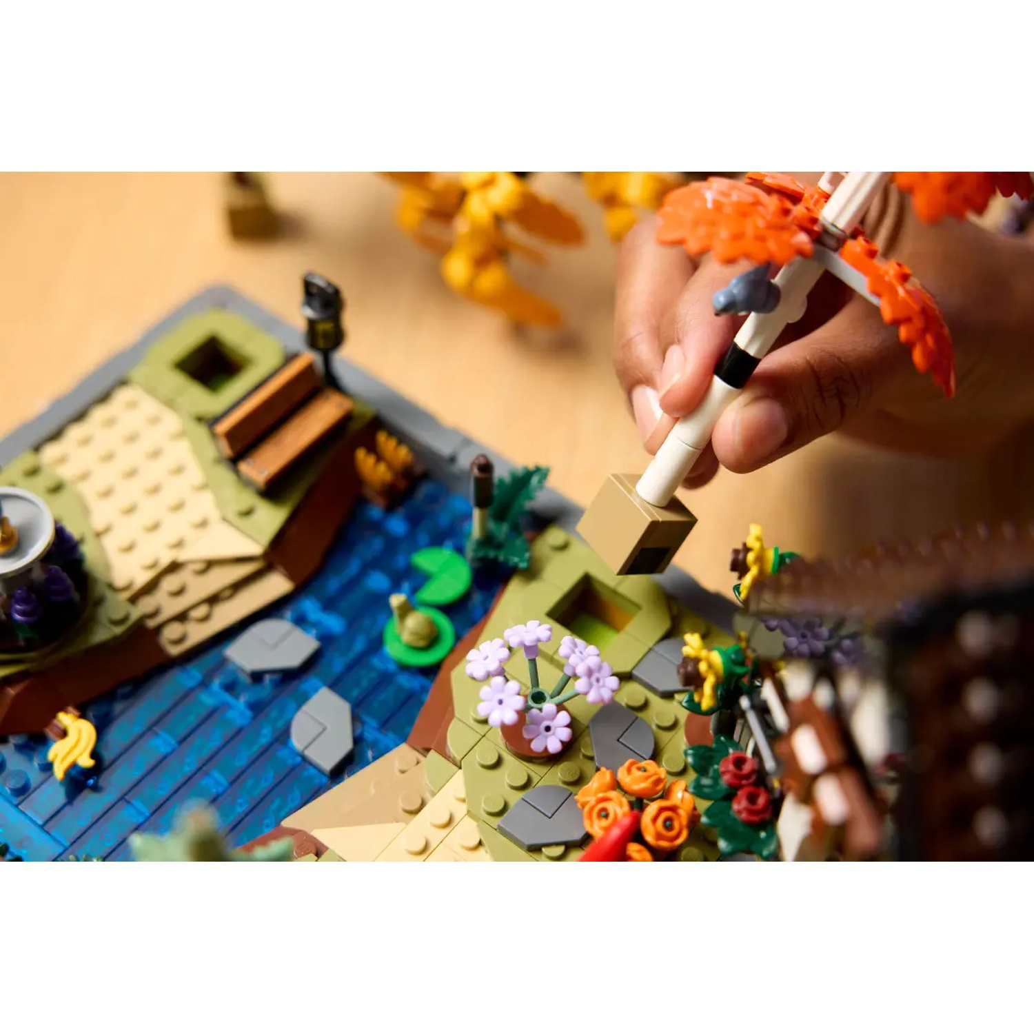 Une main tient un parapluie Lego au-dessus d'un étang Lego. L'étang a un pont, des fleurs et une petite île avec un escargot.
