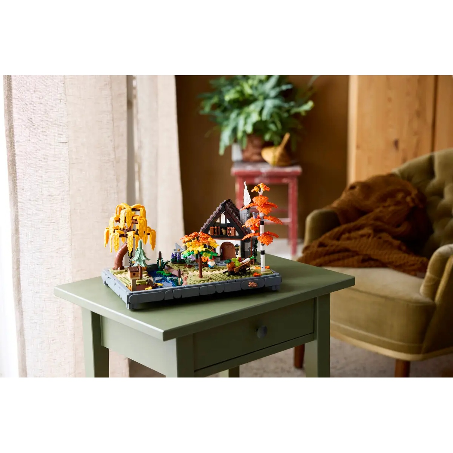 Un diorama Lego d'une maison et d'arbres sur une table verte. Une chaise verte et une plante sont en arrière-plan.