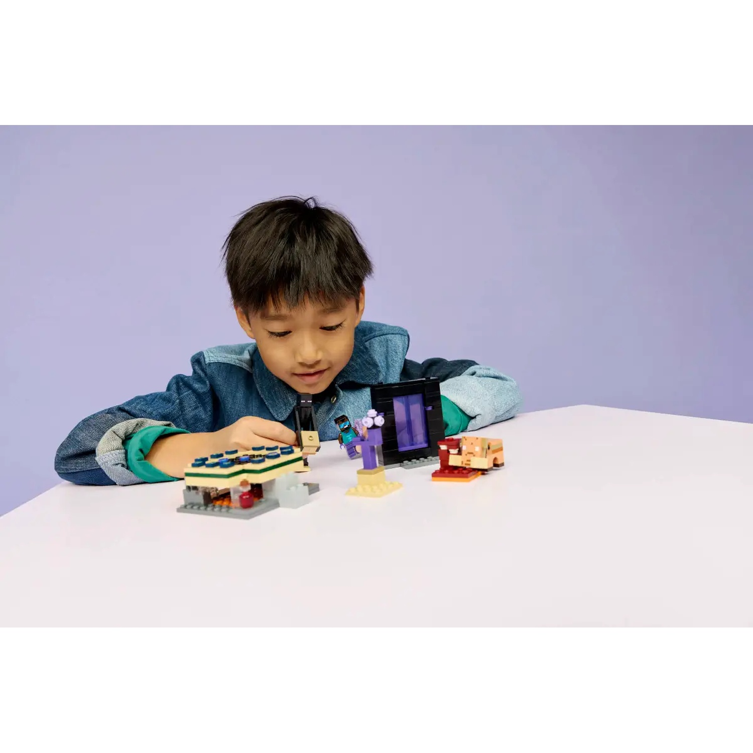 Un garçon avec une veste en jean joue avec des sets Lego Minecraft sur une table blanche. Fond violet. Les sets Lego incluent un portail et un bâtiment.