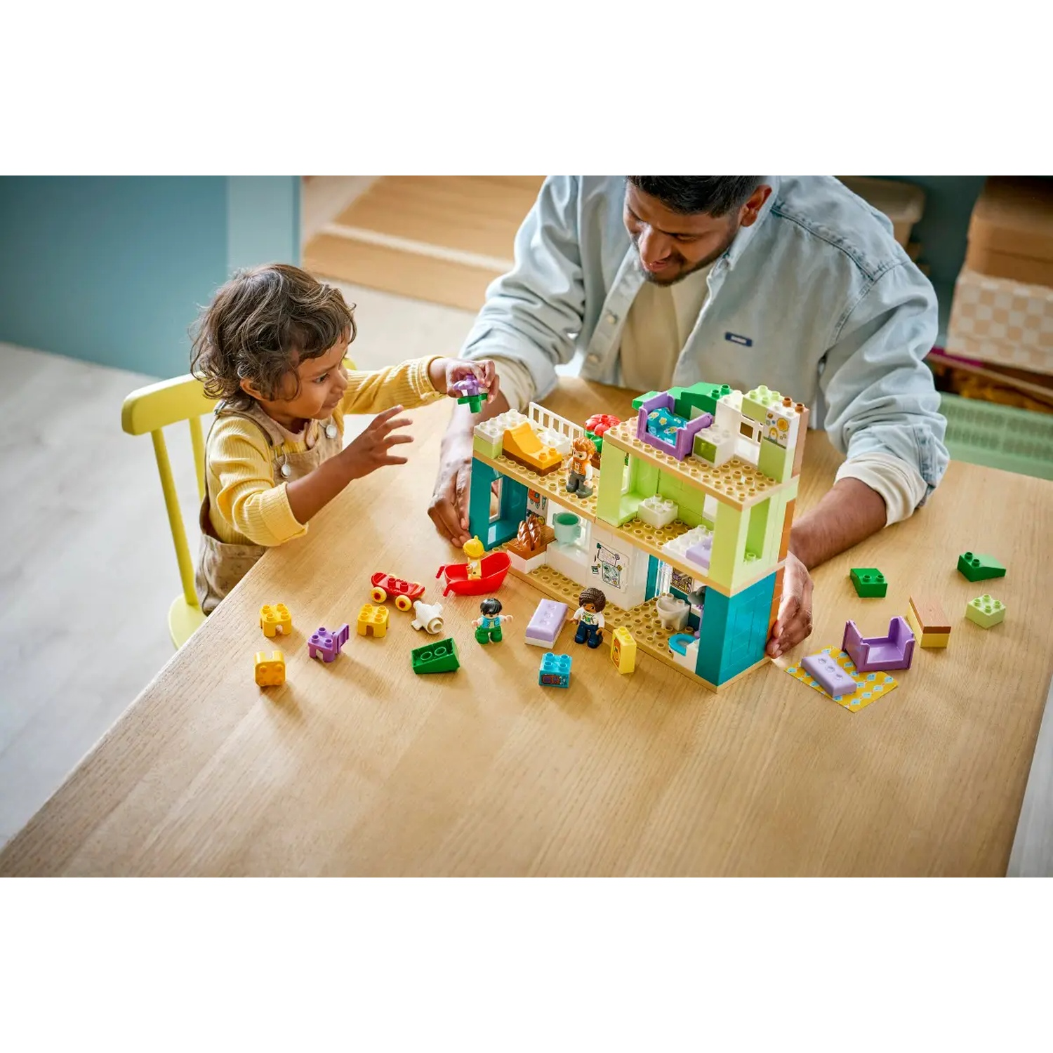 Un enfant et un adulte jouent avec un jeu de maison Lego sur une table en bois. Des jouets sont éparpillés.