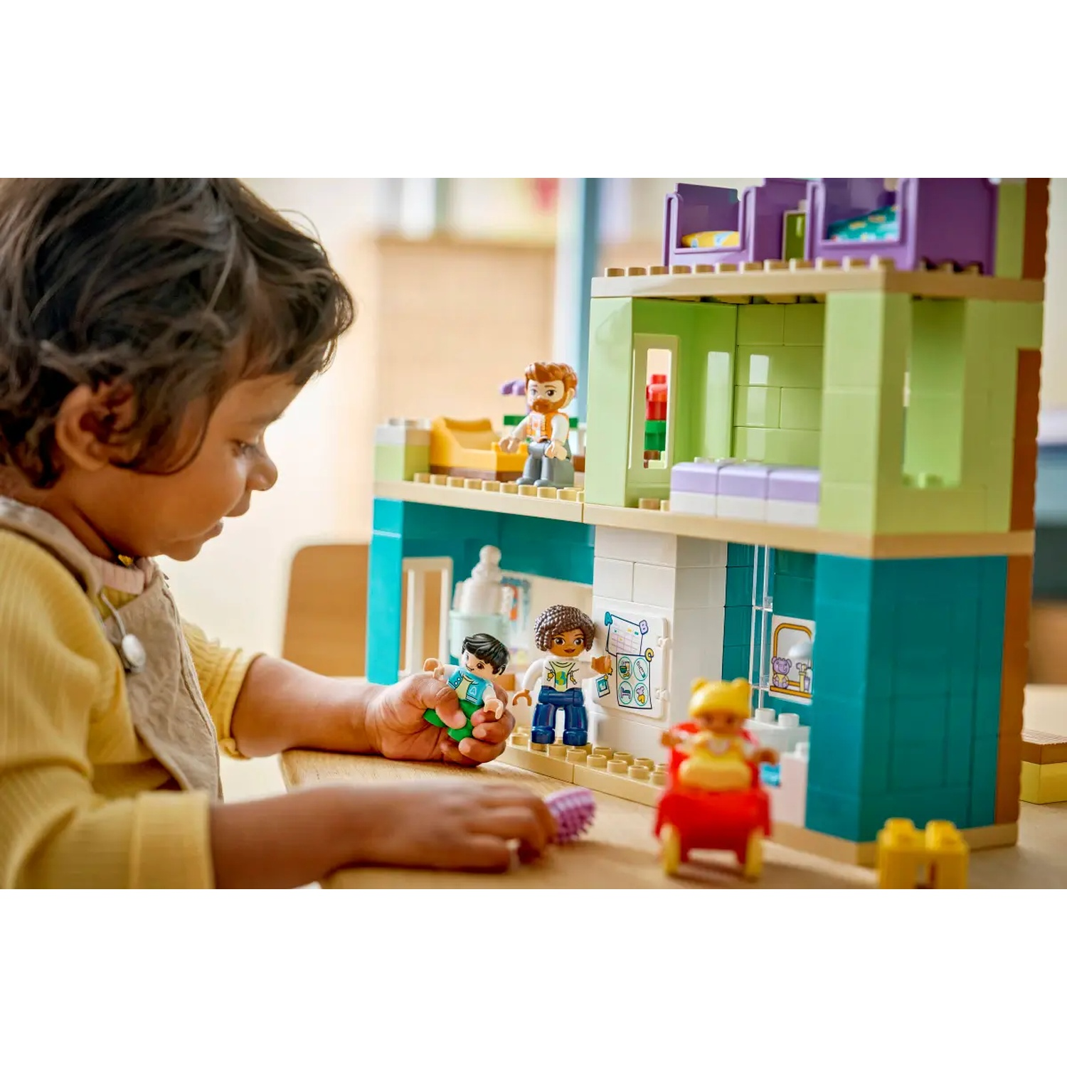 Un enfant joue avec une maison Lego. Des figurines Lego sont sur les niveaux, et l'enfant en tient une. La maison est verte, sarcelle et blanche.