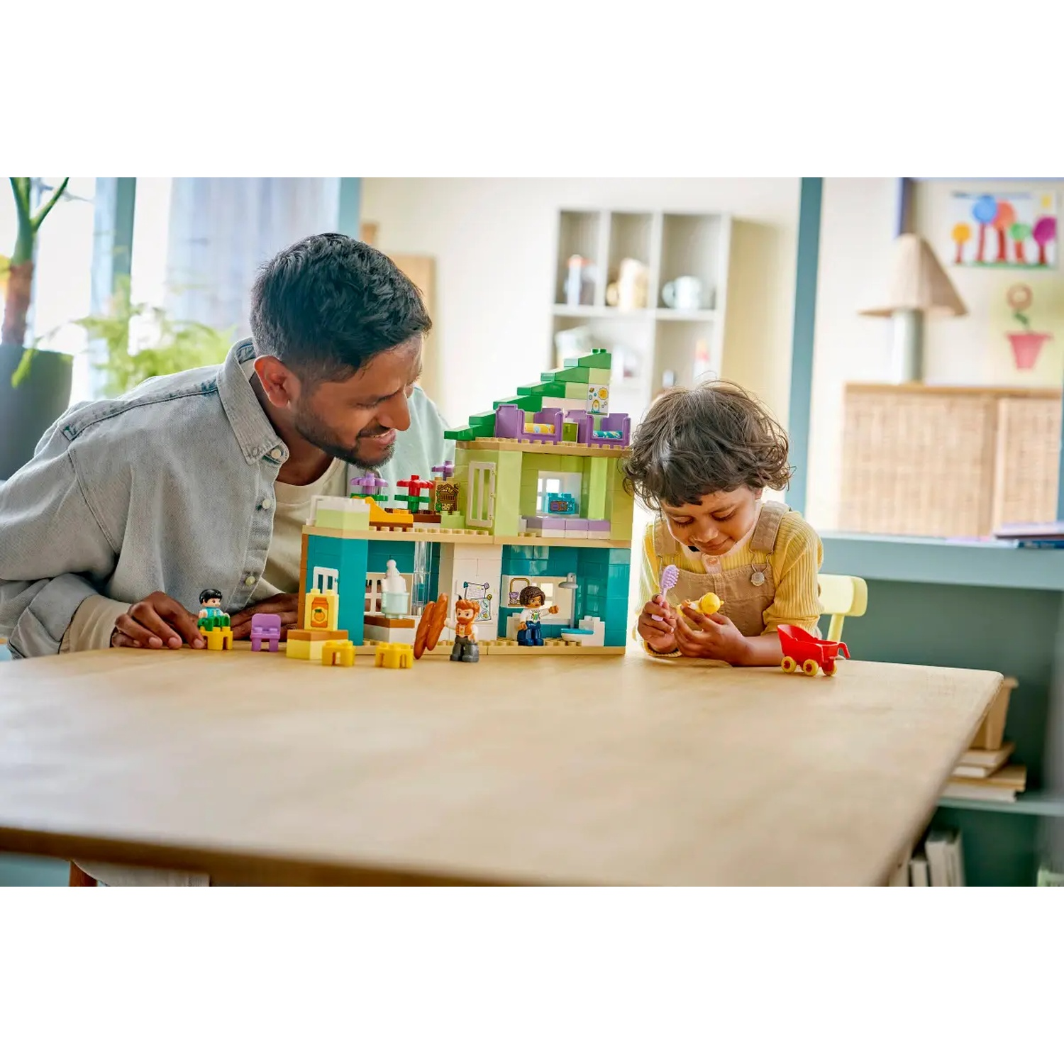 Un homme et un enfant jouent avec des Lego. Une maison en Lego est sur une table. L'enfant tient un jouet jaune.