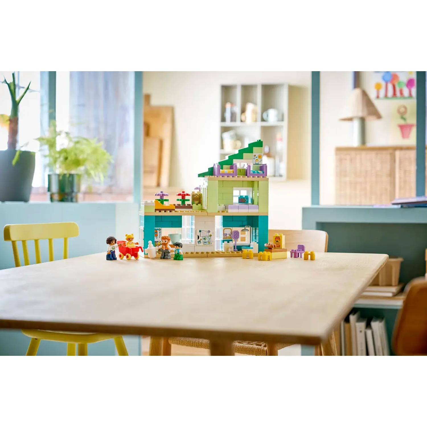 Une maison Lego avec des figurines, posée sur une table. Comprend des meubles et des plantes. Arrière-plan : une pièce avec fenêtre et étagères.