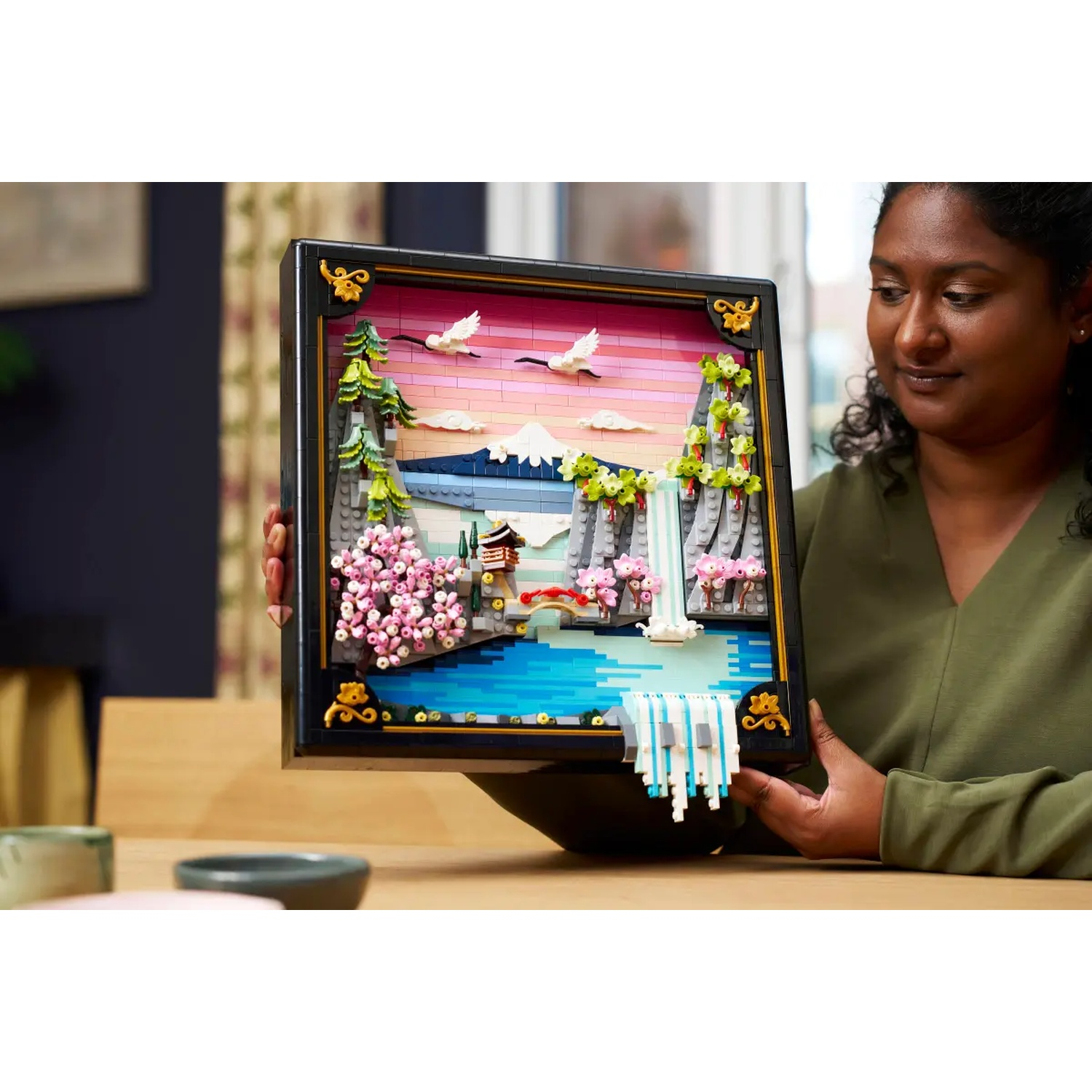 A woman holds a Lego diorama of a Japanese landscape, featuring a waterfall, cherry blossoms, and a mountain.