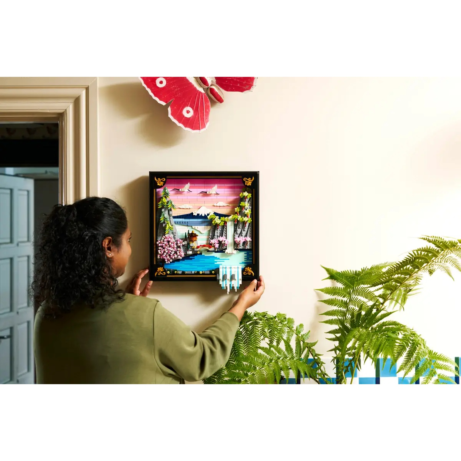 A woman hangs a framed art piece with a landscape scene. A red butterfly decorates the wall. Green ferns and plants are visible.