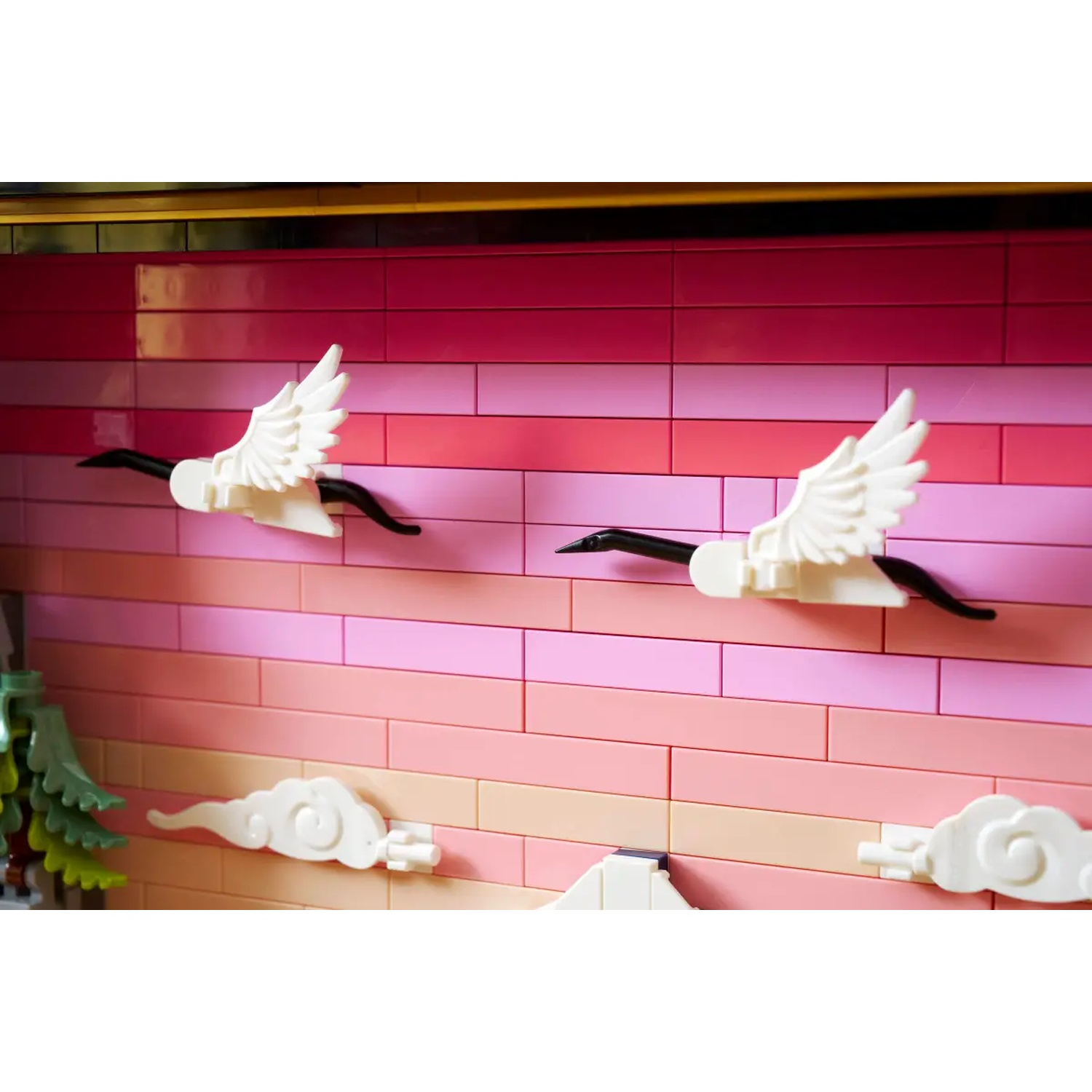 Two white cranes with black beaks and necks fly against a pink and orange brick wall. White clouds are also present.