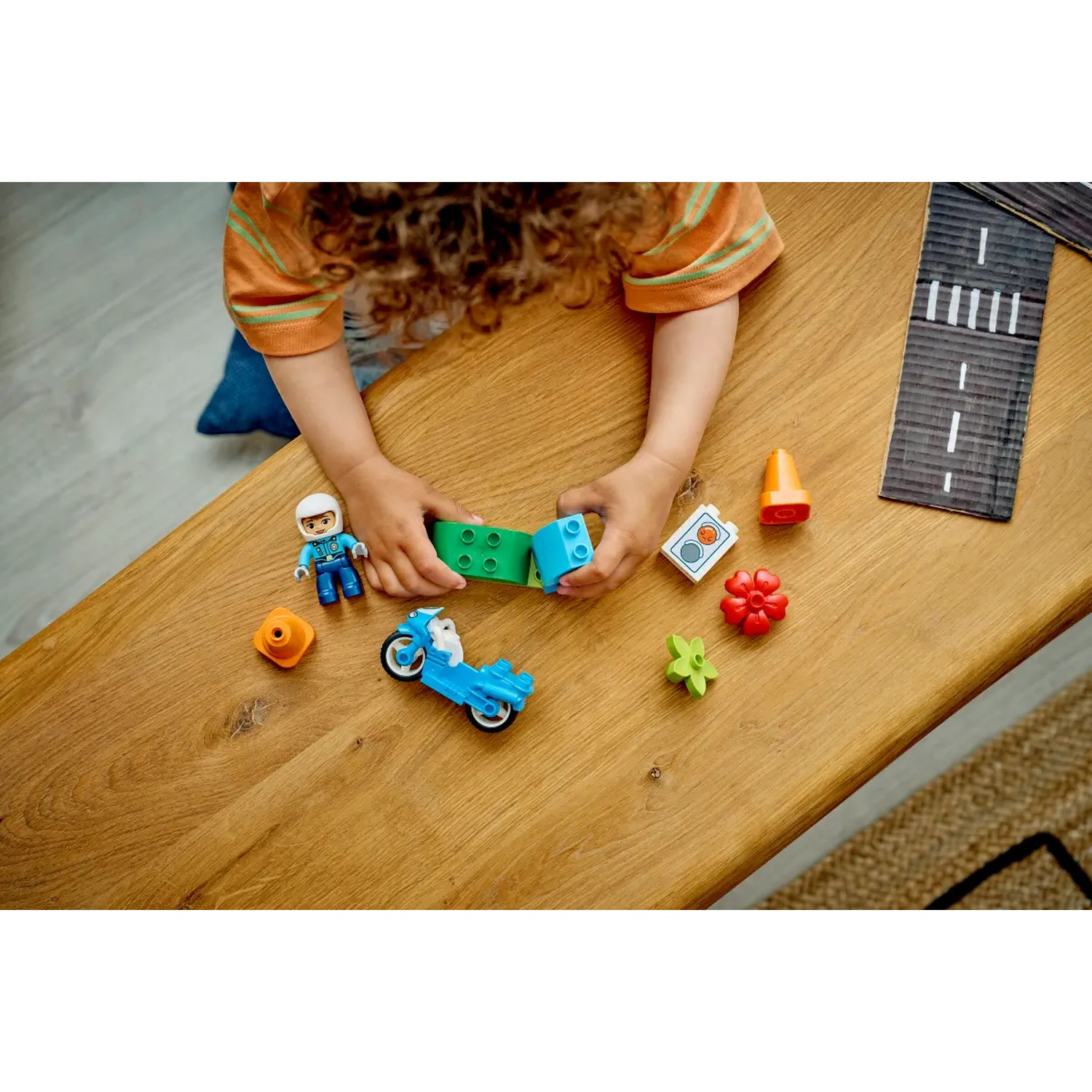 Un enfant joue avec des blocs et jouets Lego Duplo colorés sur une table en bois. Une moto bleue est visible.