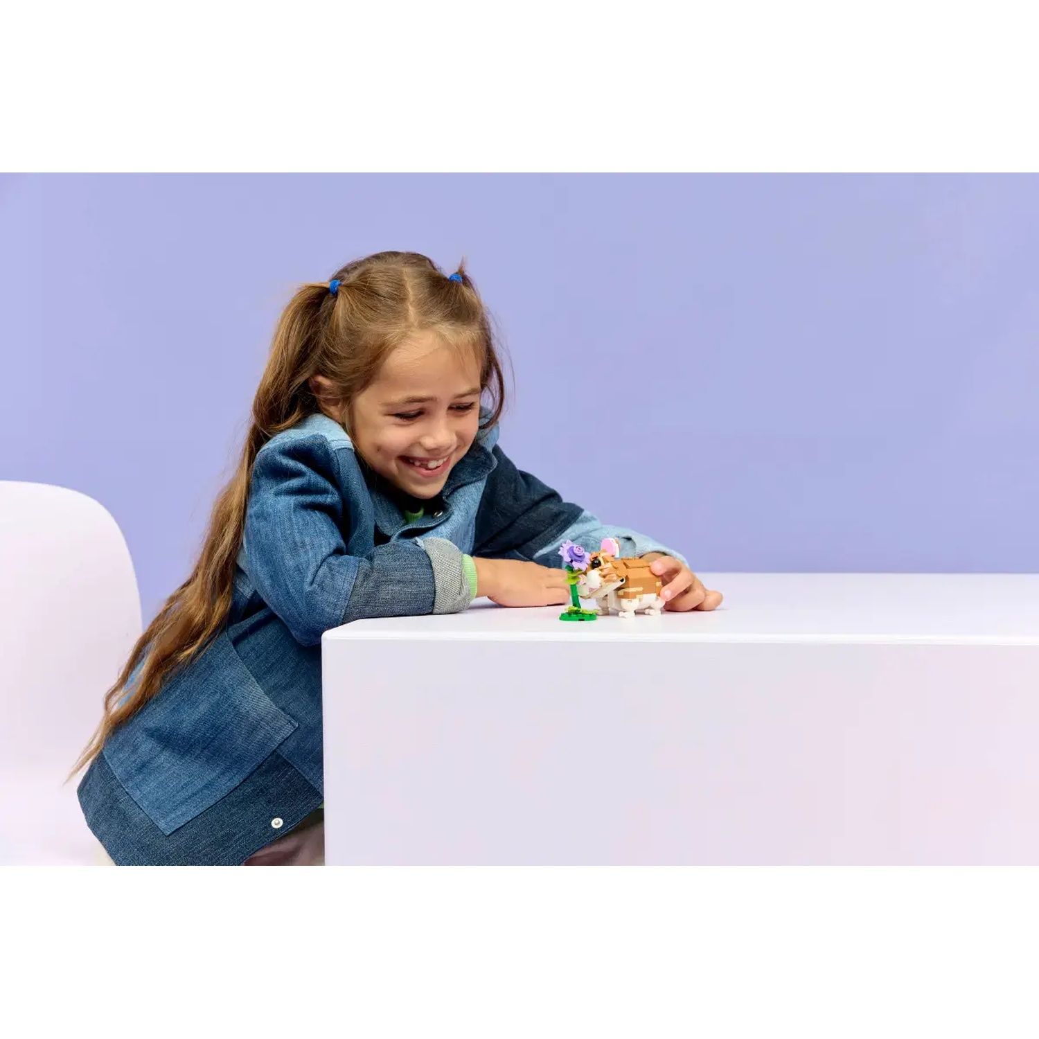 Een meisje glimlacht naar een speelgoeddier op een witte tafel. Ze draagt een spijkerjasje. De achtergrond is lichtpaars.