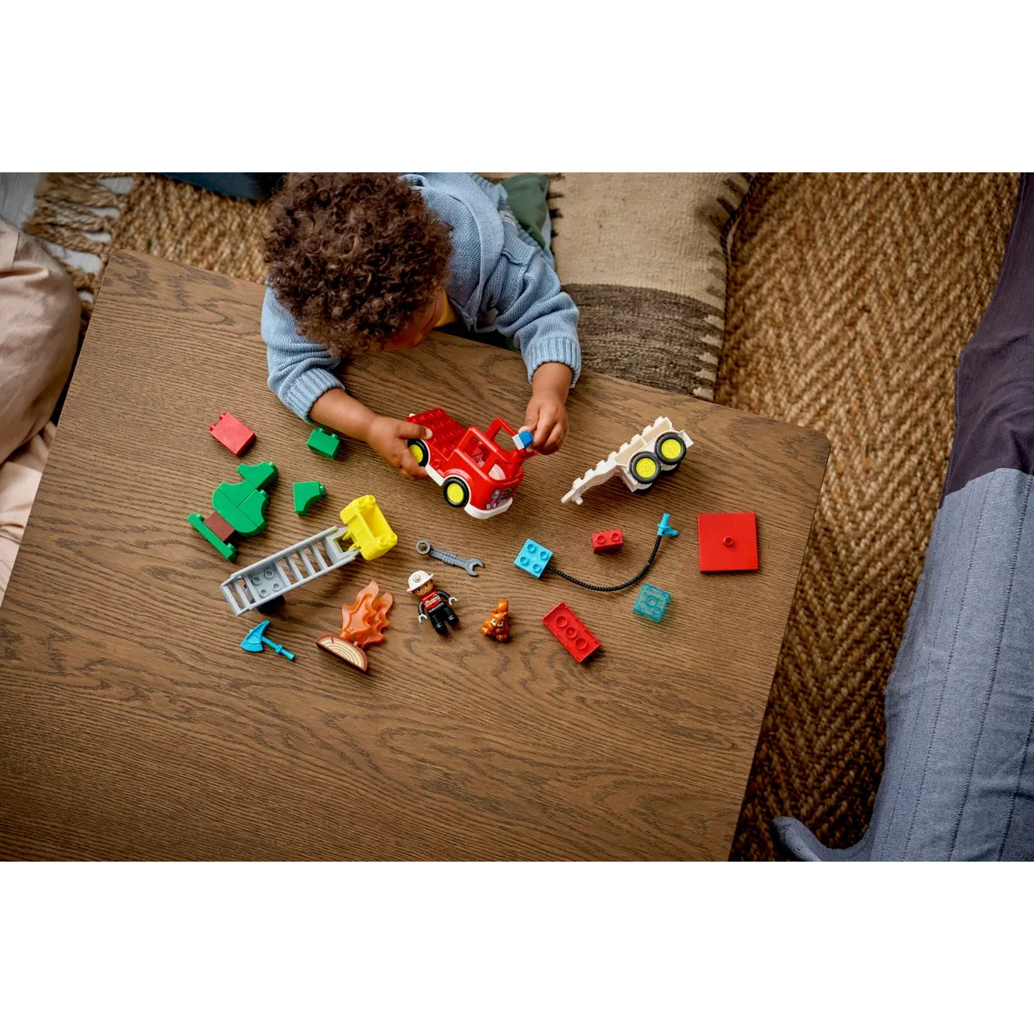 Un enfant joue avec un camion de pompier et des blocs jouets. Diverses pièces de jouets sont éparpillées sur une table en bois, devant un tapis tissé.
