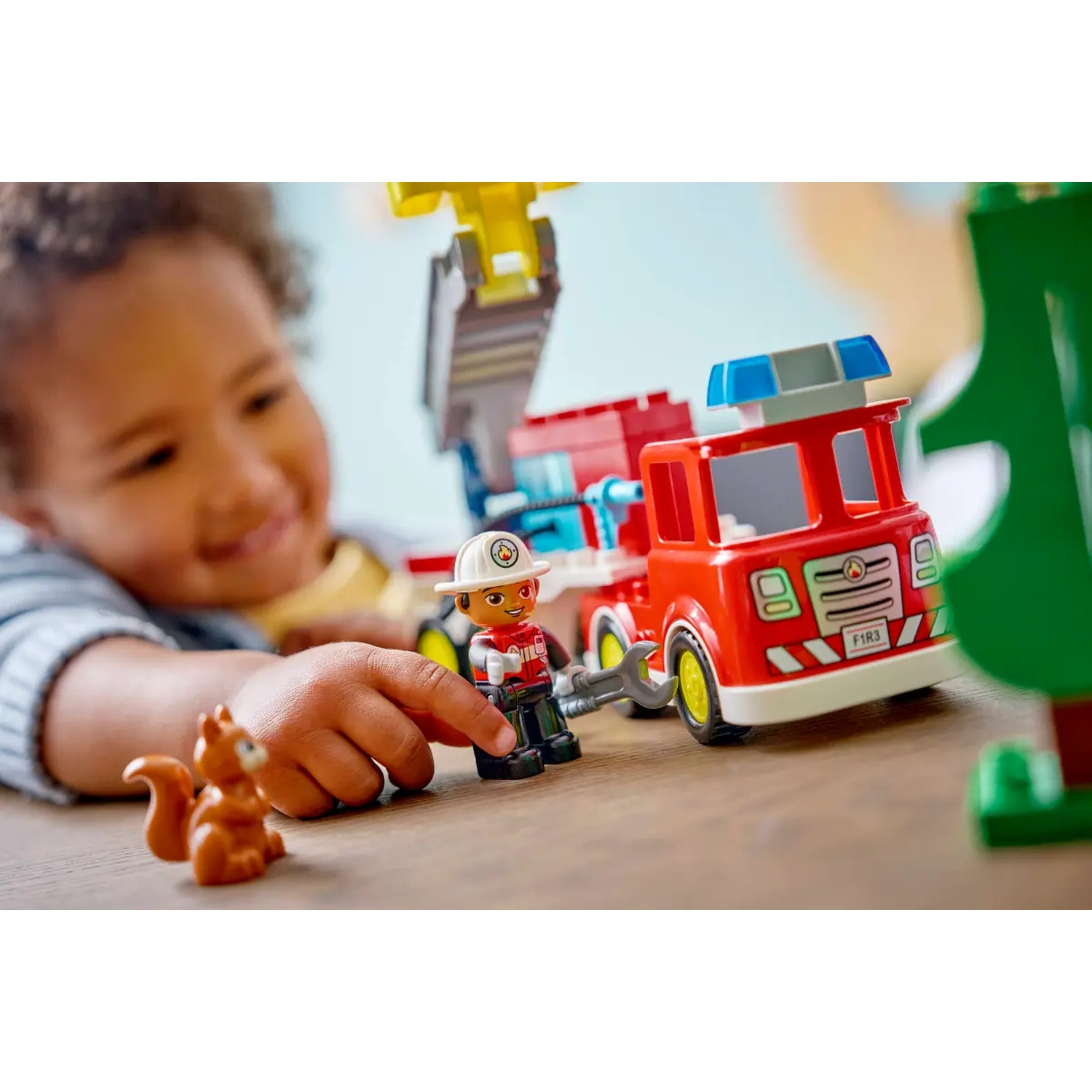Un enfant joue avec un camion de pompier rouge, une figurine de pompier et un jouet d'écureuil brun. L'enfant sourit, concentré sur les jouets.