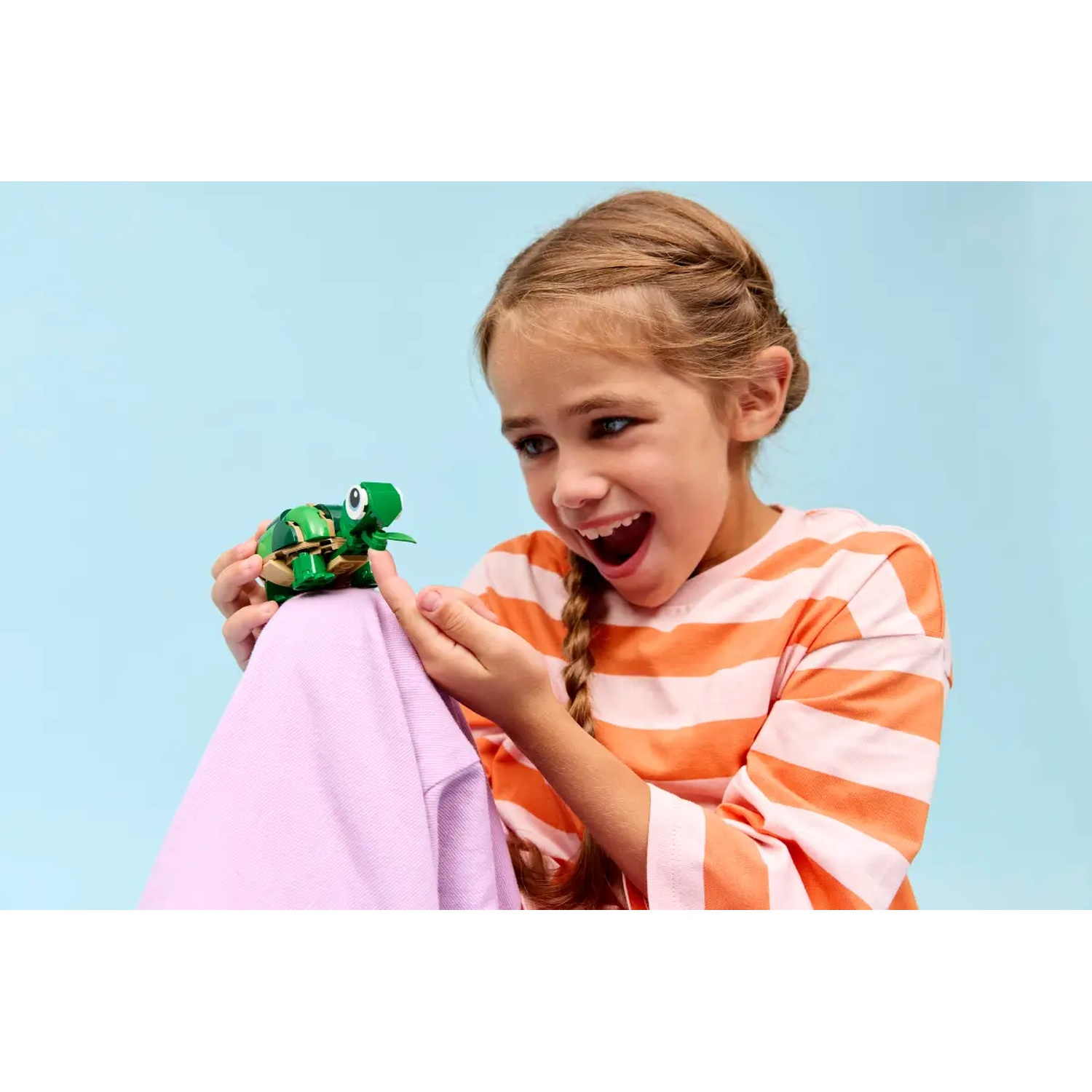 A girl smiles at a green turtle toy. She wears an orange striped shirt and holds the toy over a purple garment.