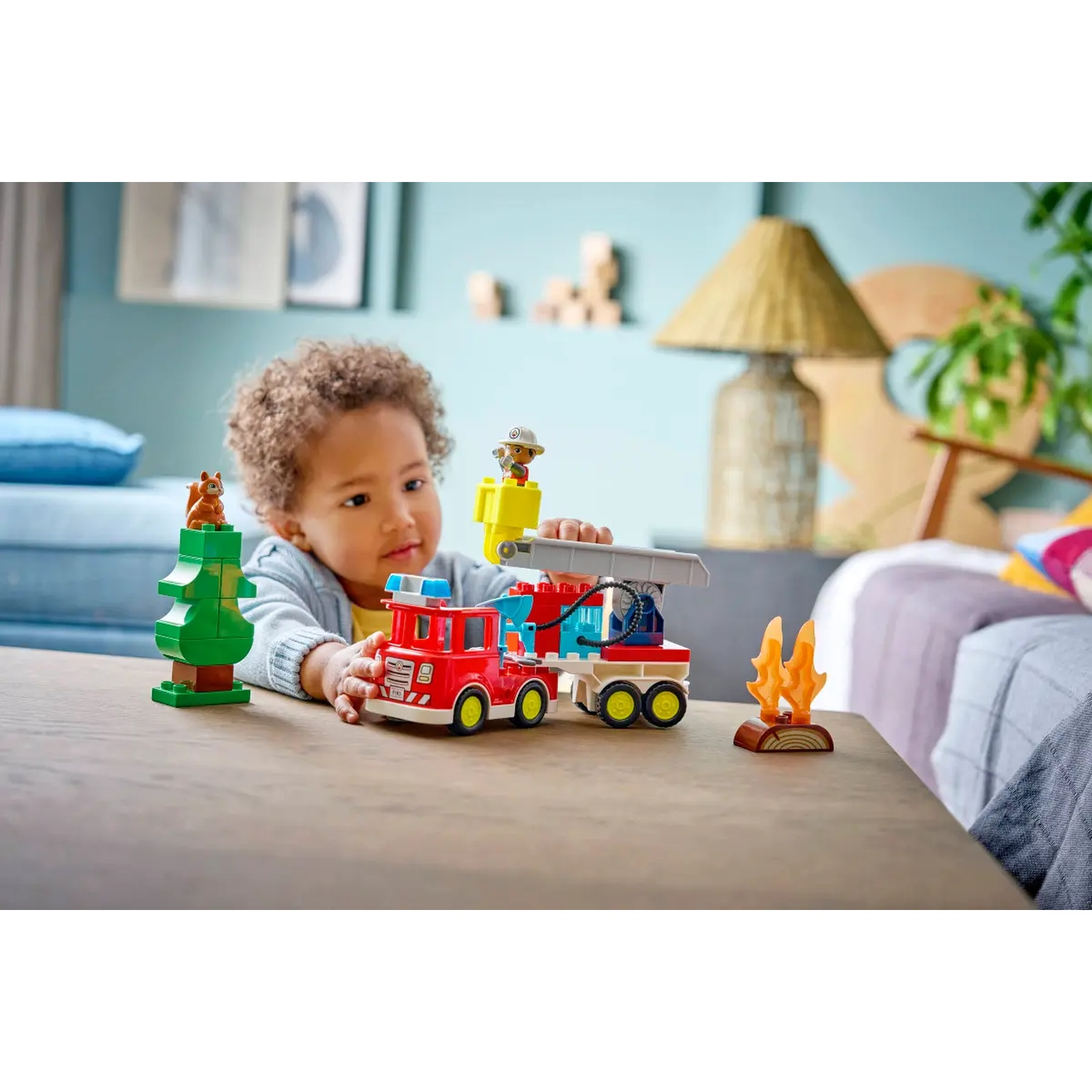 Un tout-petit joue avec un jouet de camion de pompiers. Un arbre et une figurine d'écureuil sont à gauche. Un feu et un pompier sont également présents.