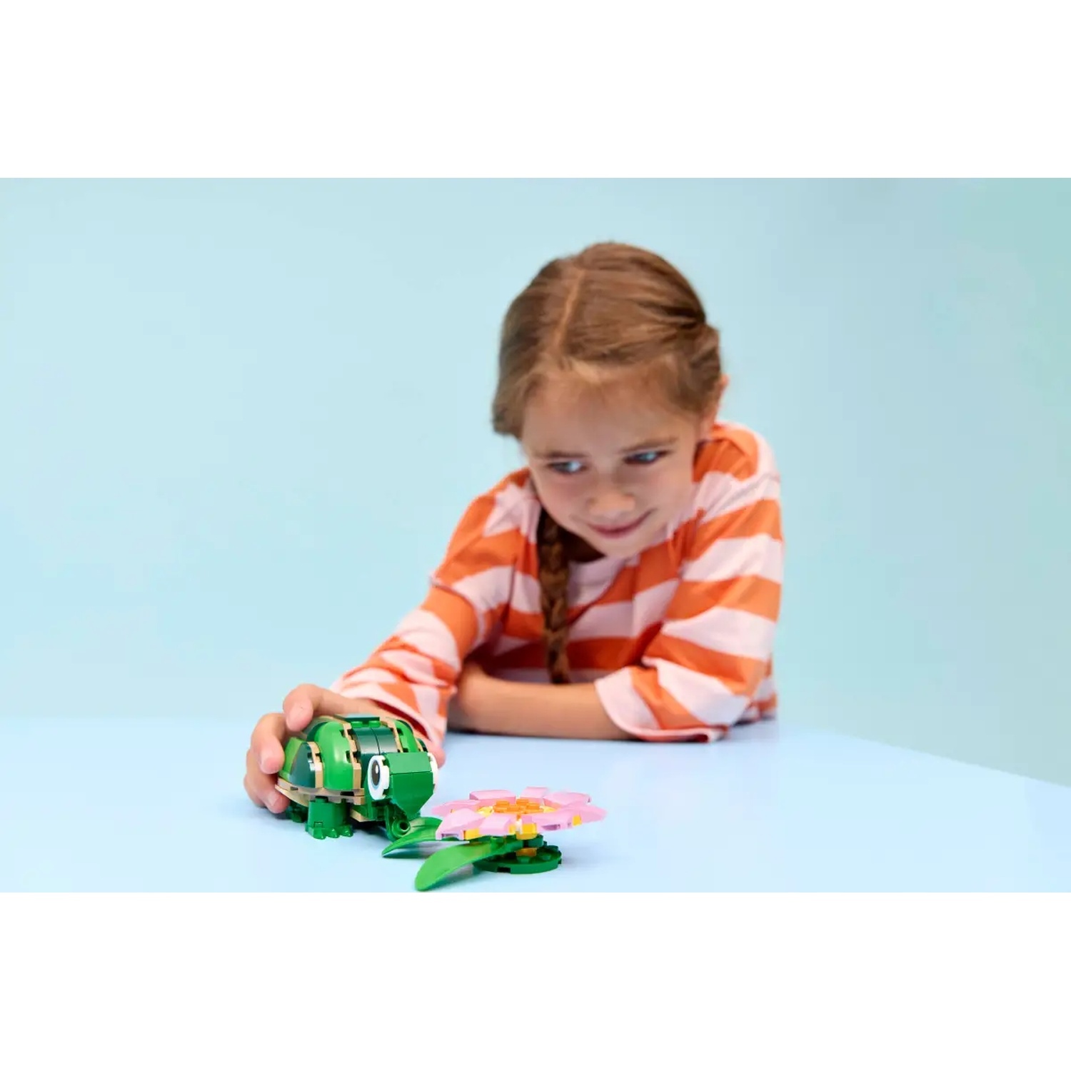 A girl looks at a green Lego frog and a flower on a blue table. She wears a striped orange shirt.