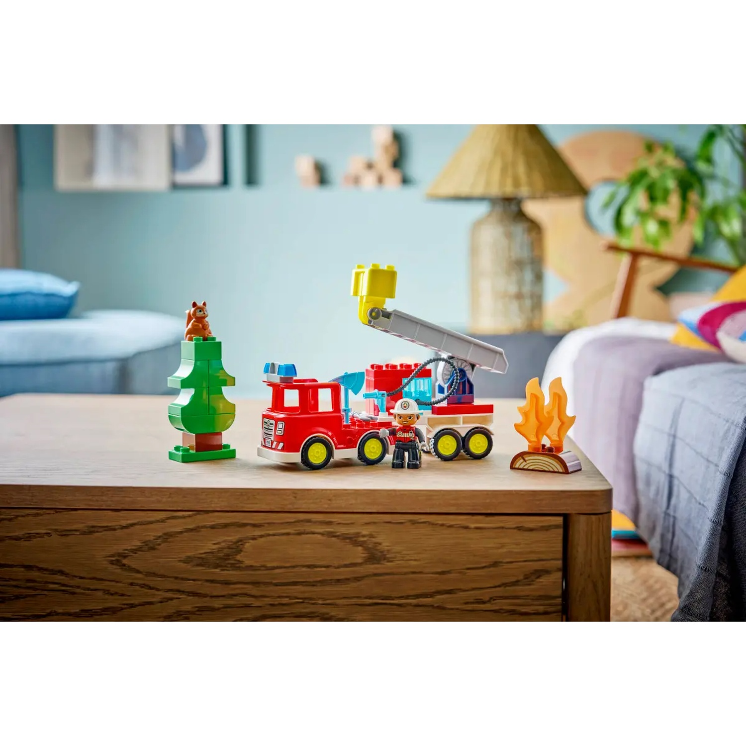 Un camion de pompiers Lego, un arbre avec un écureuil et une figurine de pompier sont sur une table en bois. Un feu est également montré. L'arrière-plan est un salon.