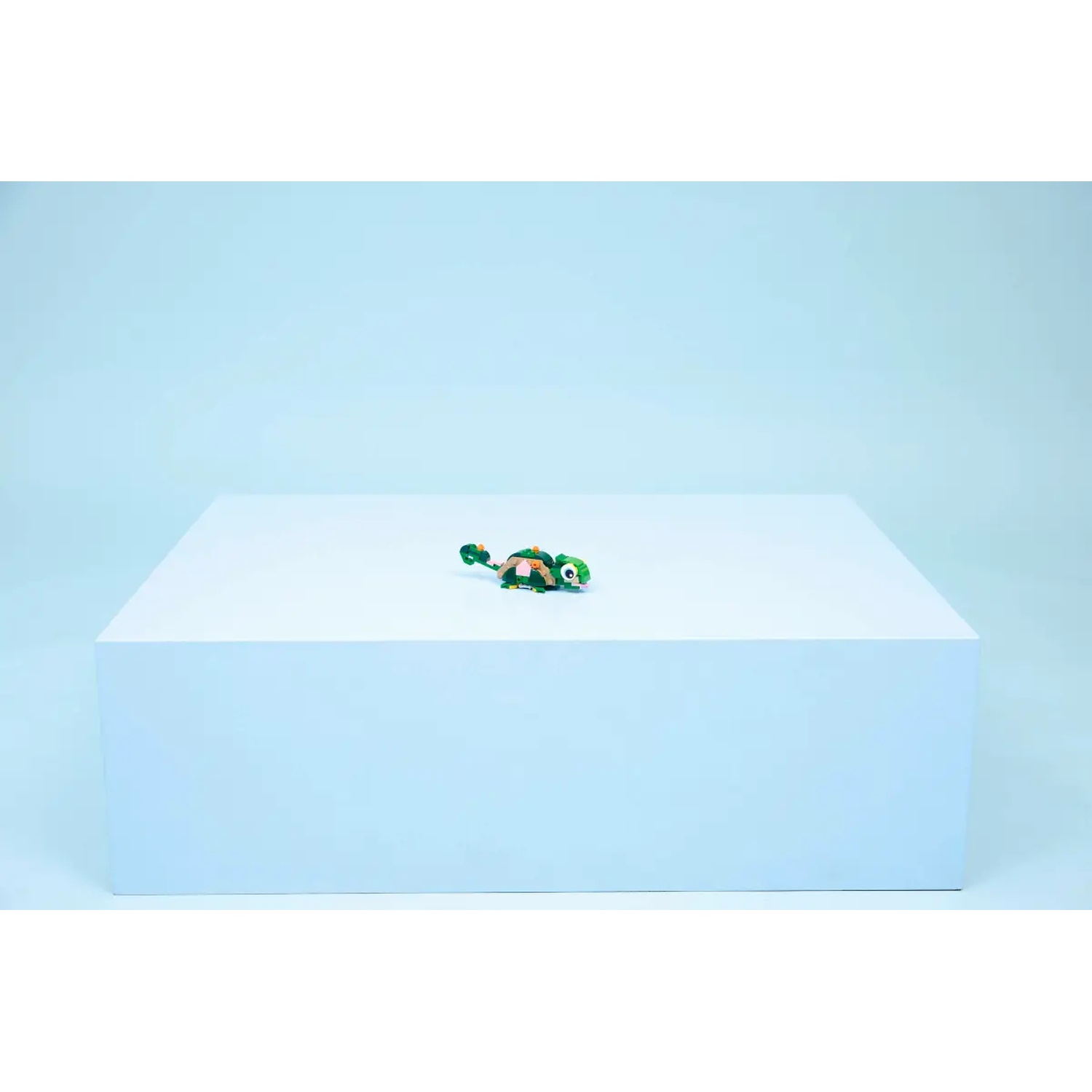 A toy turtle sits on a white cube, against a light blue background. The turtle is green, orange and black.