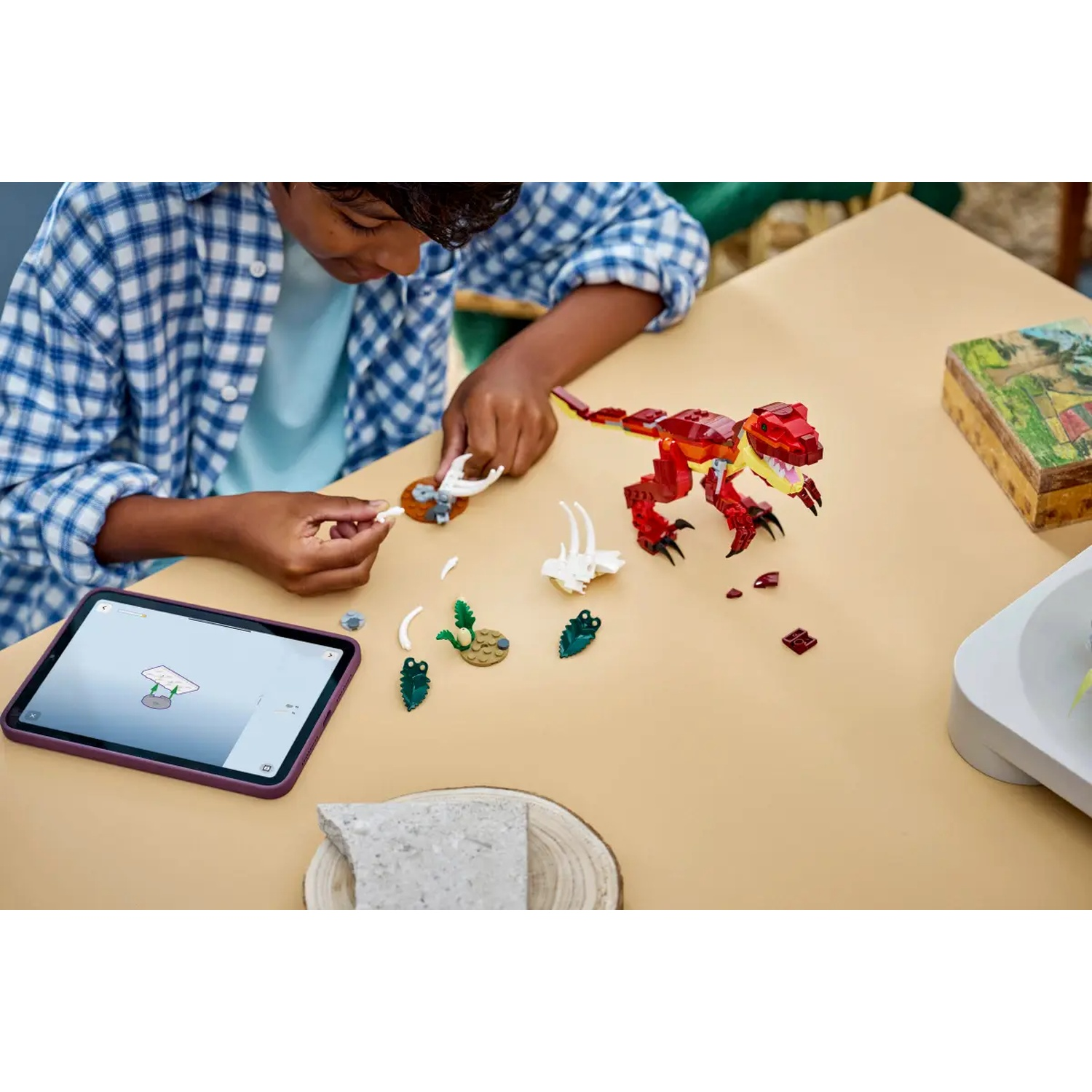 Een kind assembleert een Lego-dinosaurus op een tafel. Een tablet toont instructies. Verschillende Lego-onderdelen en een houten doos liggen in de buurt.