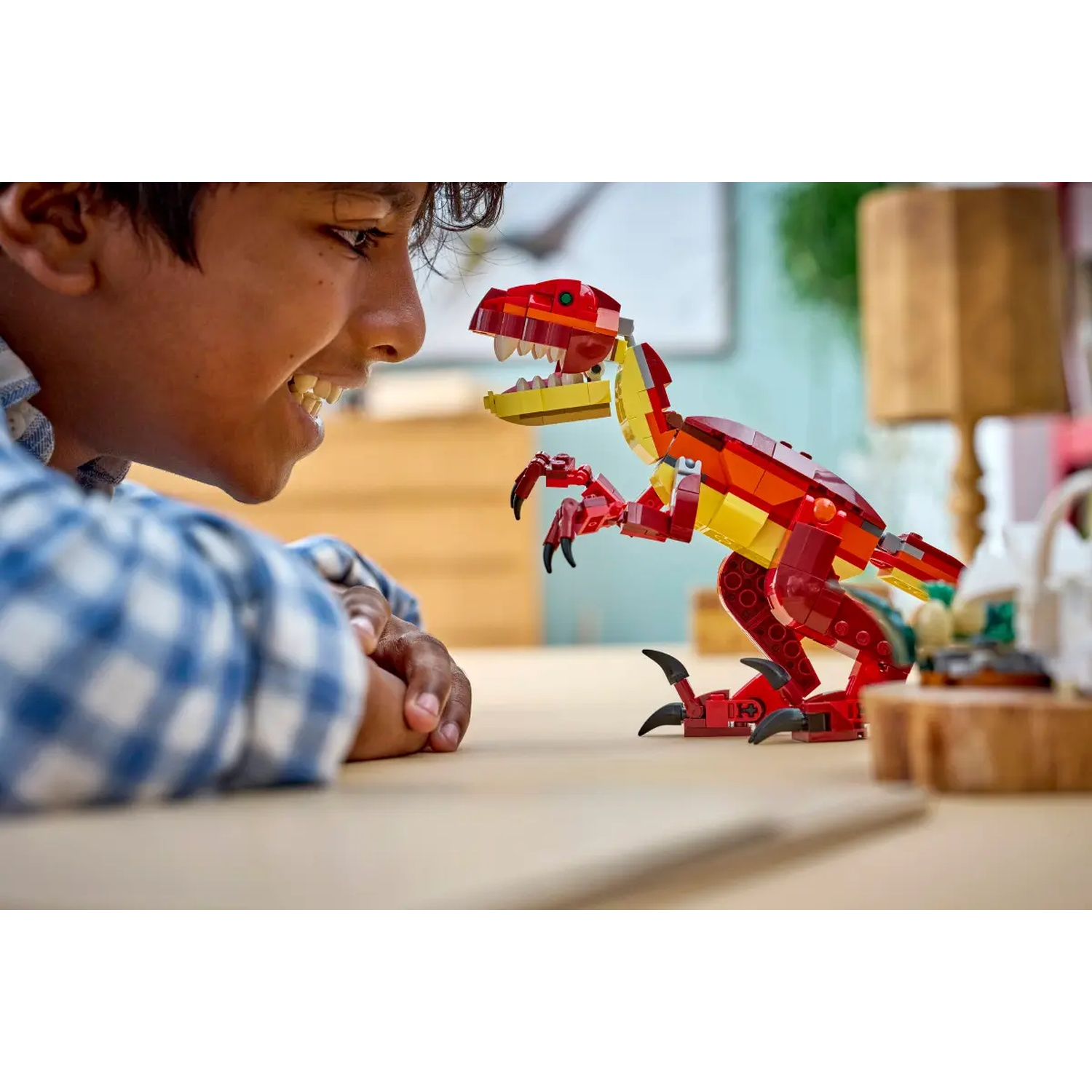 Een lachende jongen kijkt naar een rode en gele Lego-dinosaurus op een houten tafel. Achtergrond heeft groene planten en een houten blok.