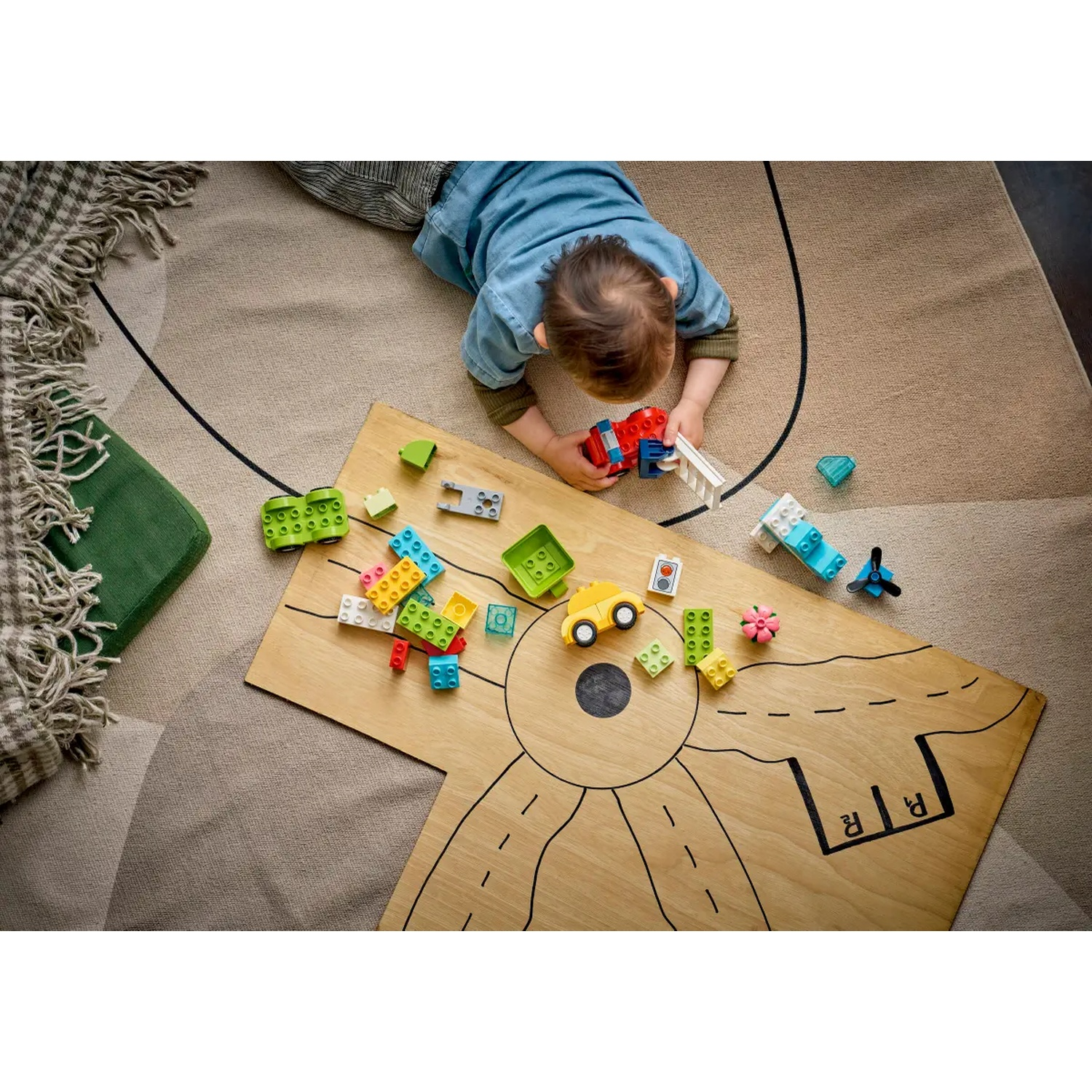 Un enfant joue avec des blocs colorés et des voitures jouets sur une planche en bois. La planche a des marquages routiers, et l'enfant est sur un tapis.