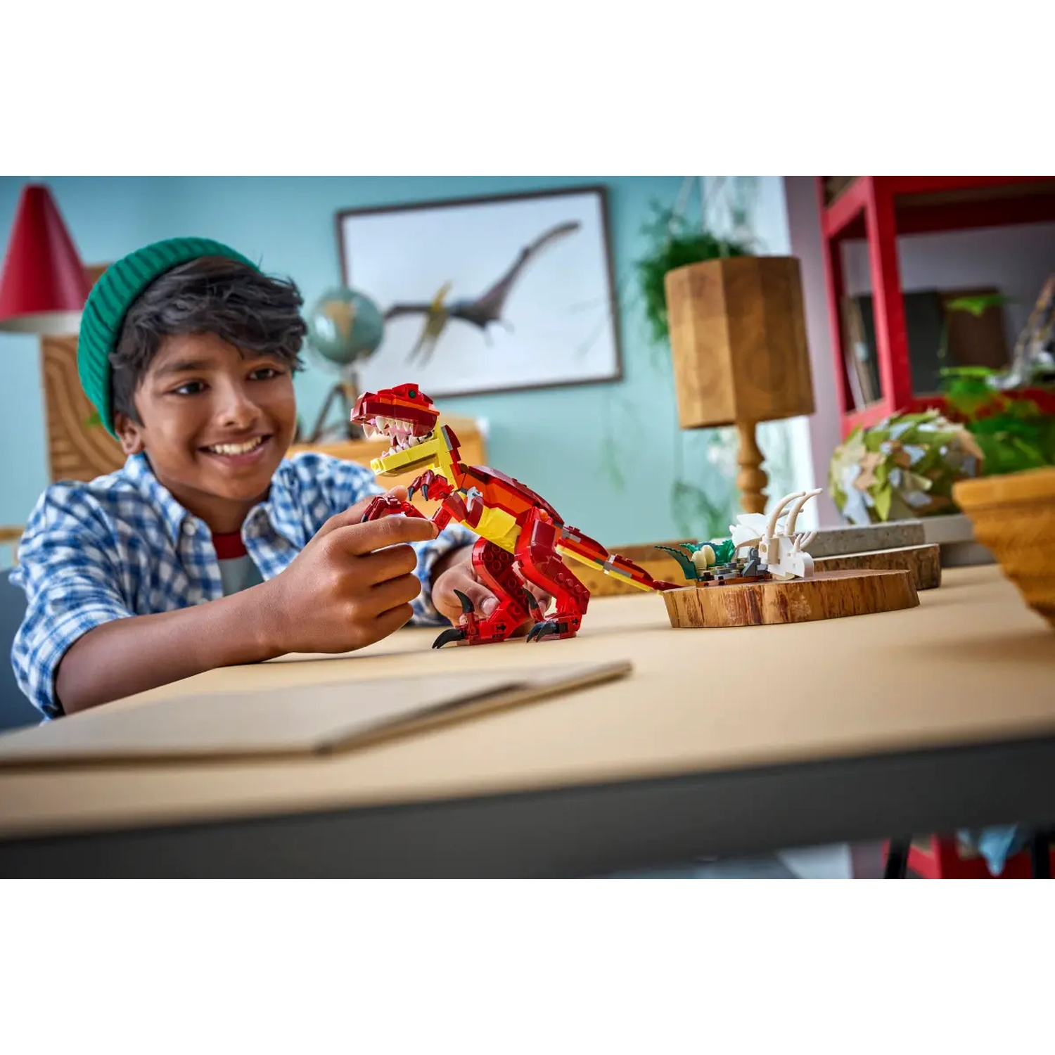 Een lachende jongen met een groene muts speelt met een rode Lego-dinosaurus op een tafel. Andere Lego-modellen zijn in de buurt.