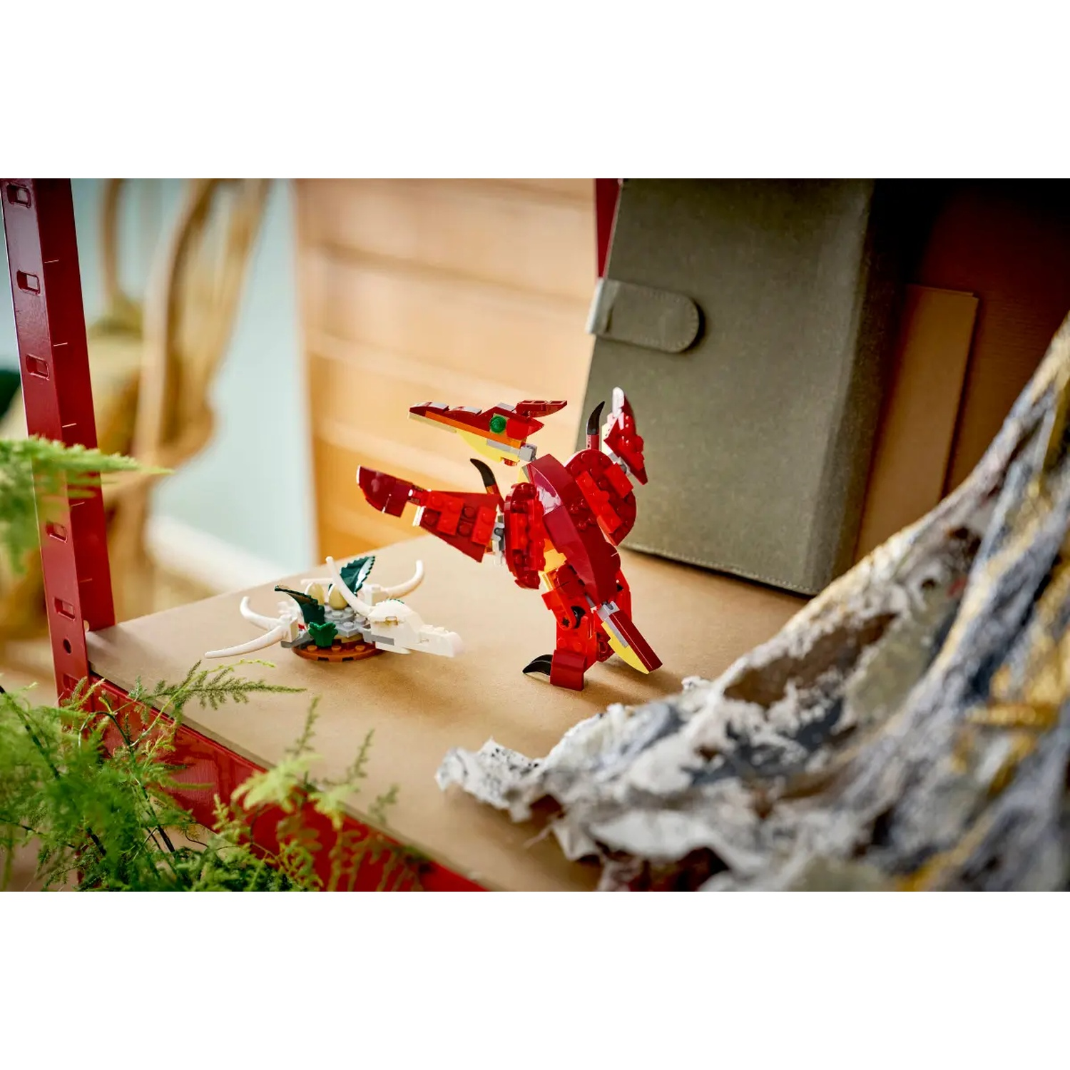 Een rode Lego-draak staat op een plank naast een witte Lego-draak. Een groene plant en een grijs boek staan op de achtergrond.