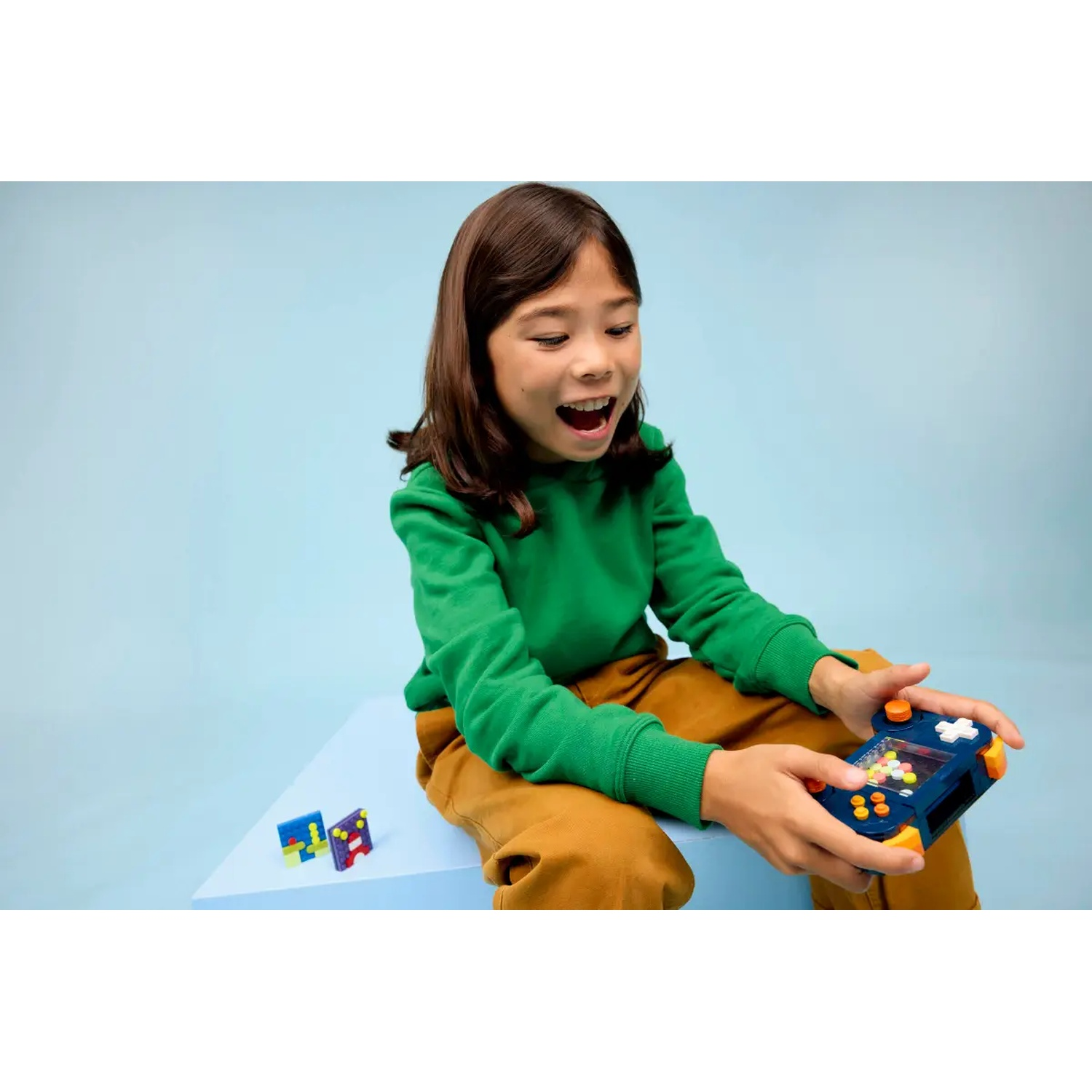 A girl is playing a game. She wears green sweater and brown pants. The game is blue and orange. Blue background.