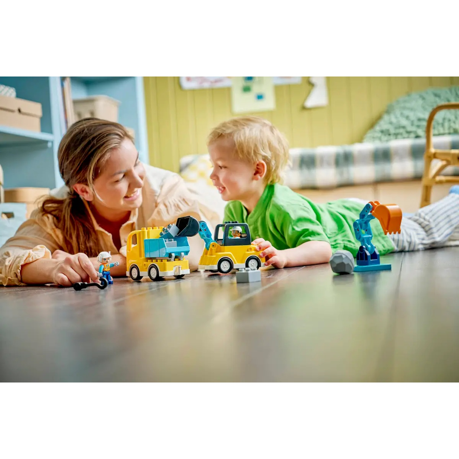 Une femme et un enfant jouent avec des véhicules de construction jouets sur un sol en bois, se souriant.